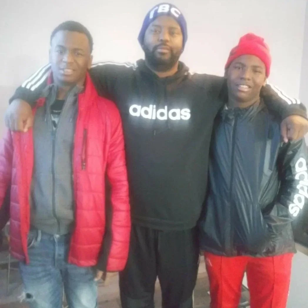 Three men standing together with their arms around each other, smiling. They are wearing casual jackets, hoodies, and beanies, and are indoors against a plain wall.