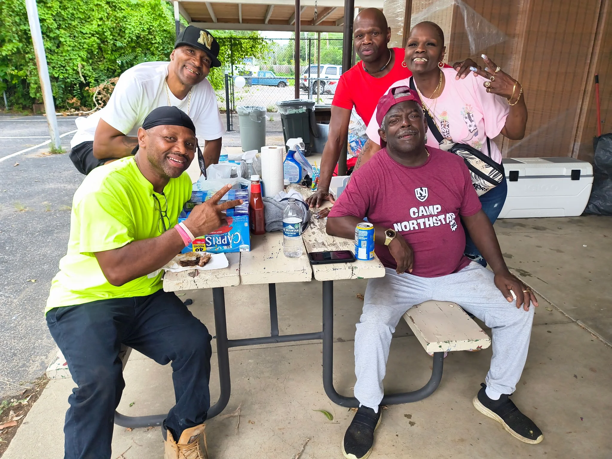 Group of six people enjoying a casual outdoor gathering around a table with food and drinks, some making peace signs and smiling.