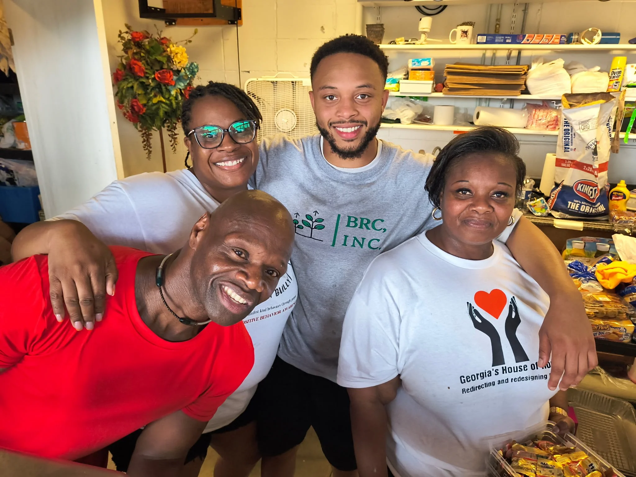 Four people smiling and posing together inside a room, with shelves and snacks in the background. One person wearing a red shirt is leaning in, while others are standing behind and beside them.