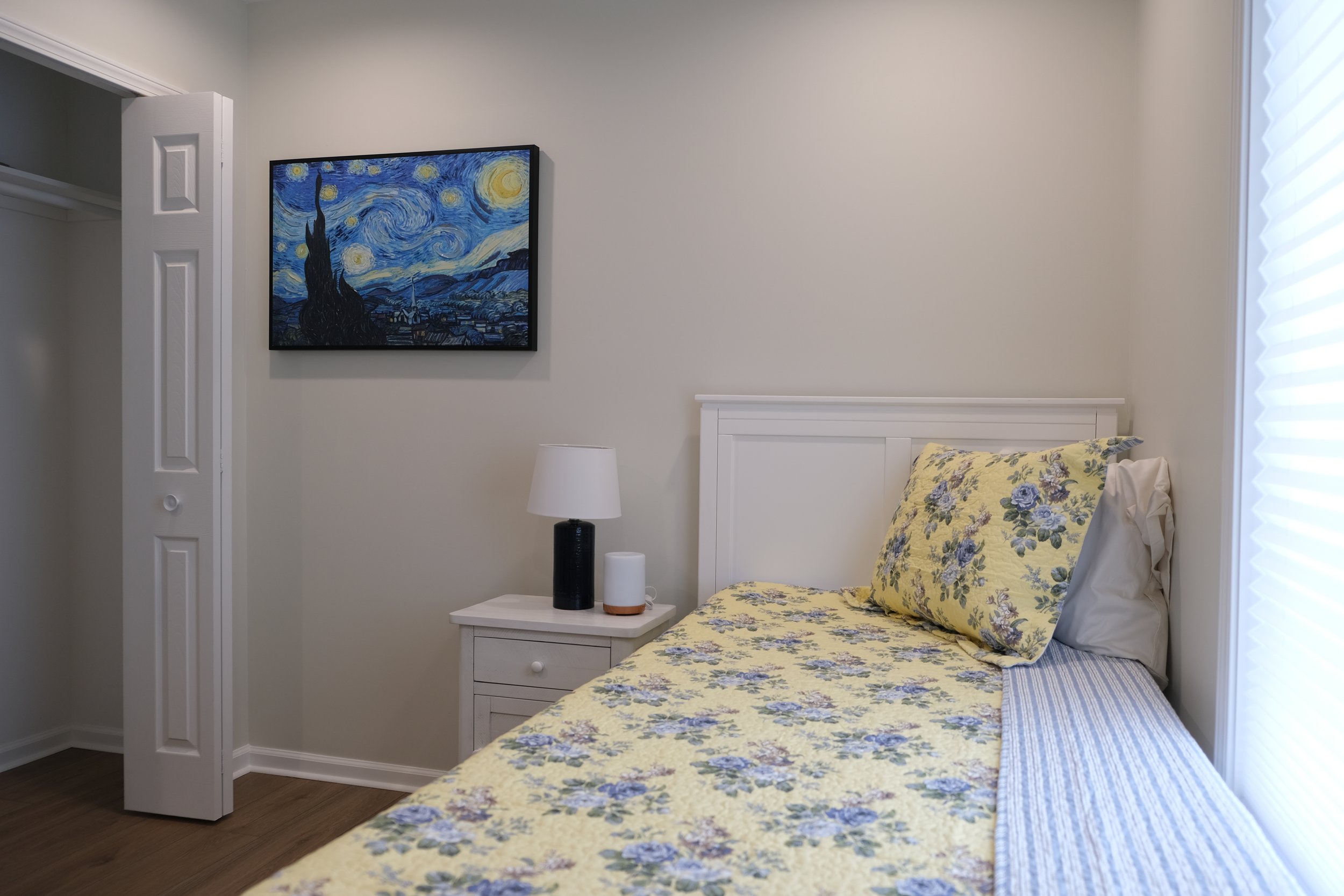 A bedroom with a white bed frame, yellow floral bedding, and a matching pillow. A nightstand with a black and white lamp and a white speaker sits beside the bed. A painting of Van Gogh's Starry Night hangs on the wall.