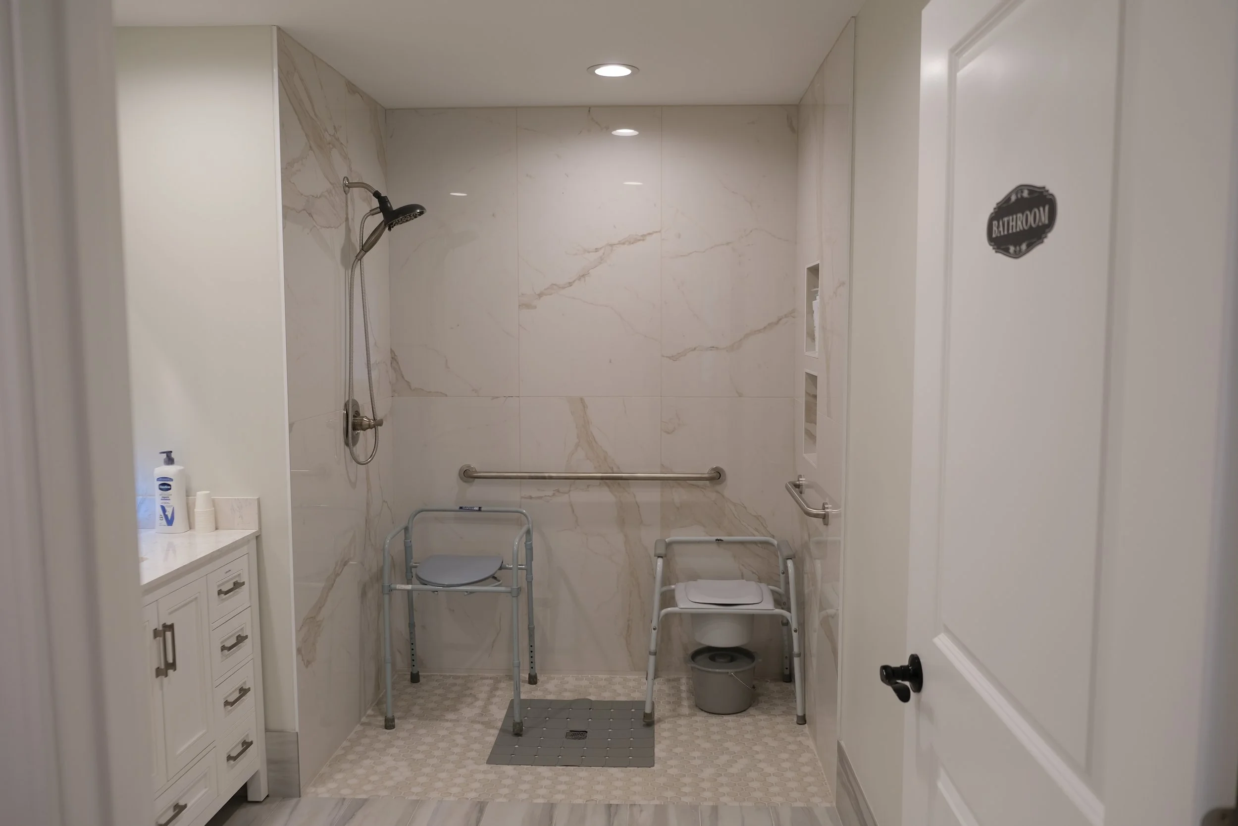 Accessible bathroom with a walk-in shower, grab bars, and shower chairs. White cabinetry, marble tiles, and a door with a "Bathroom" sign.