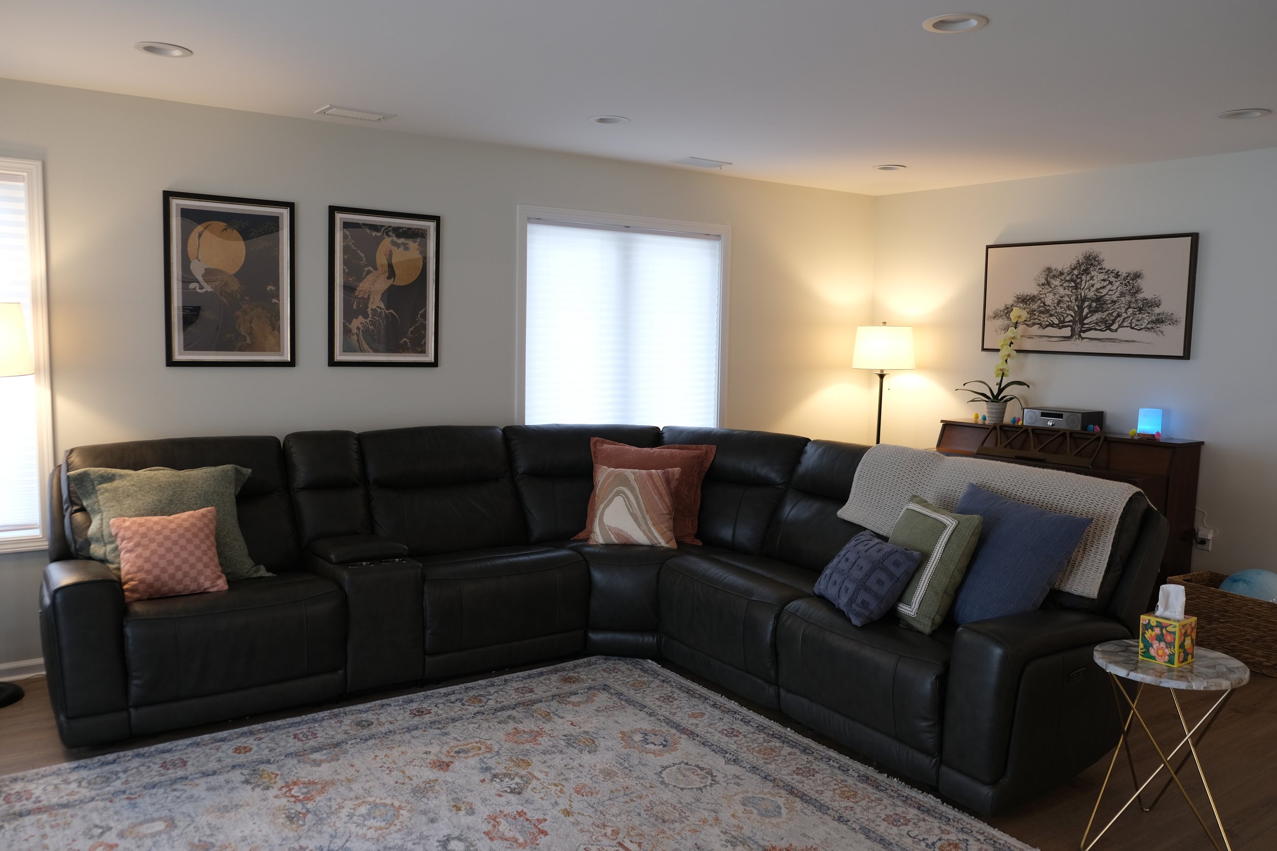 Living room with a black leather sectional sofa, colorful throw pillows, a patterned area rug, a side table with tissues, a potted orchid, a floor lamp, and framed artwork on the walls.
