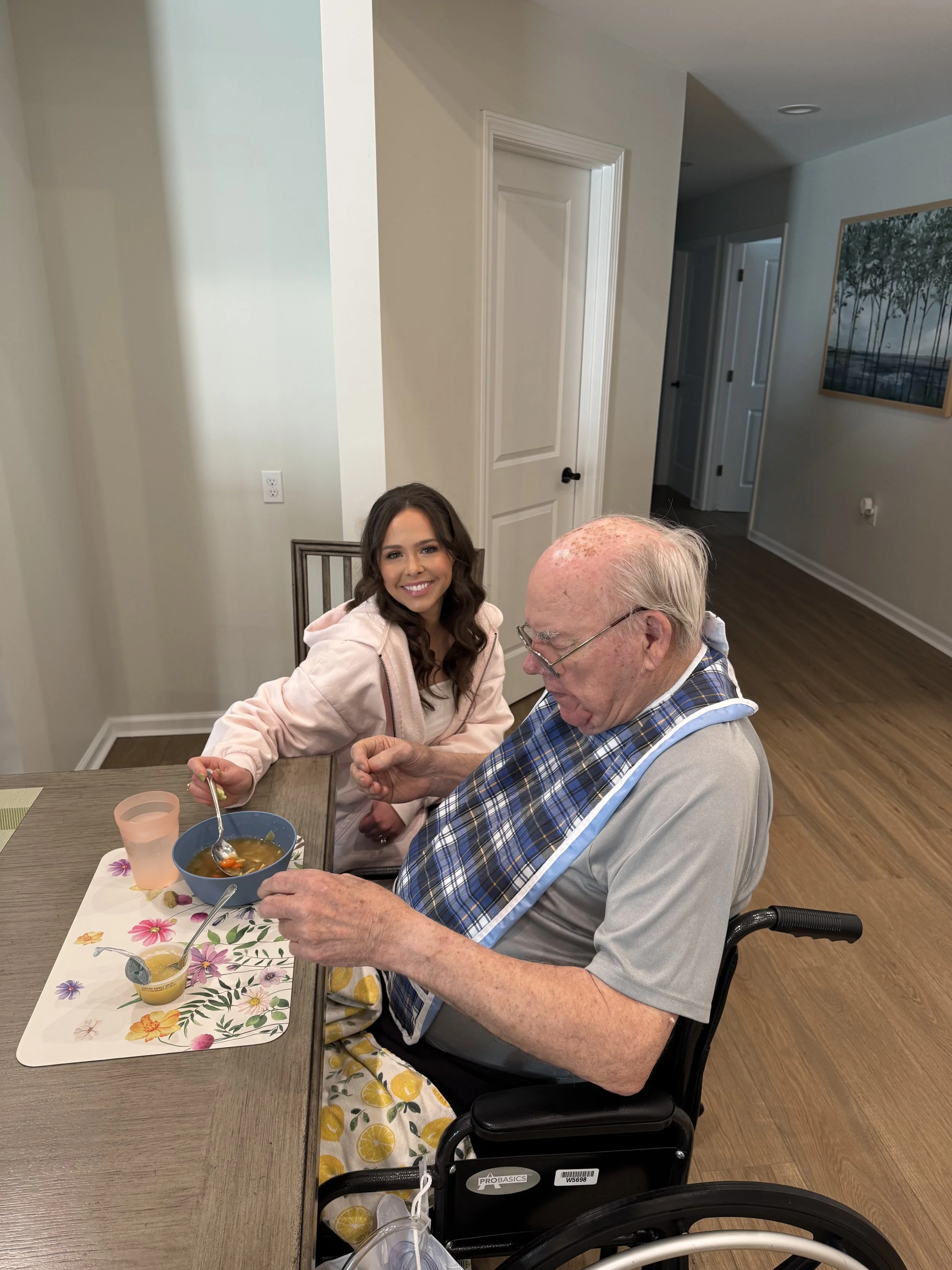 A young woman sitting beside an elderly man in a wheelchair, sharing a meal at a table, smiling at the camera in a bright, home interior.
