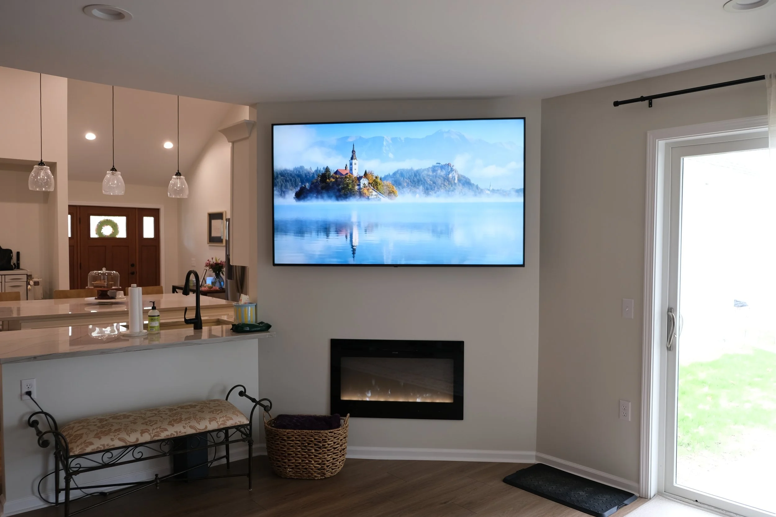 Living room with a wall-mounted TV displaying a scenic lake view with a church on a small island, fireplace below the TV, sliding glass door to the right, and kitchen with pendant lights in the background.