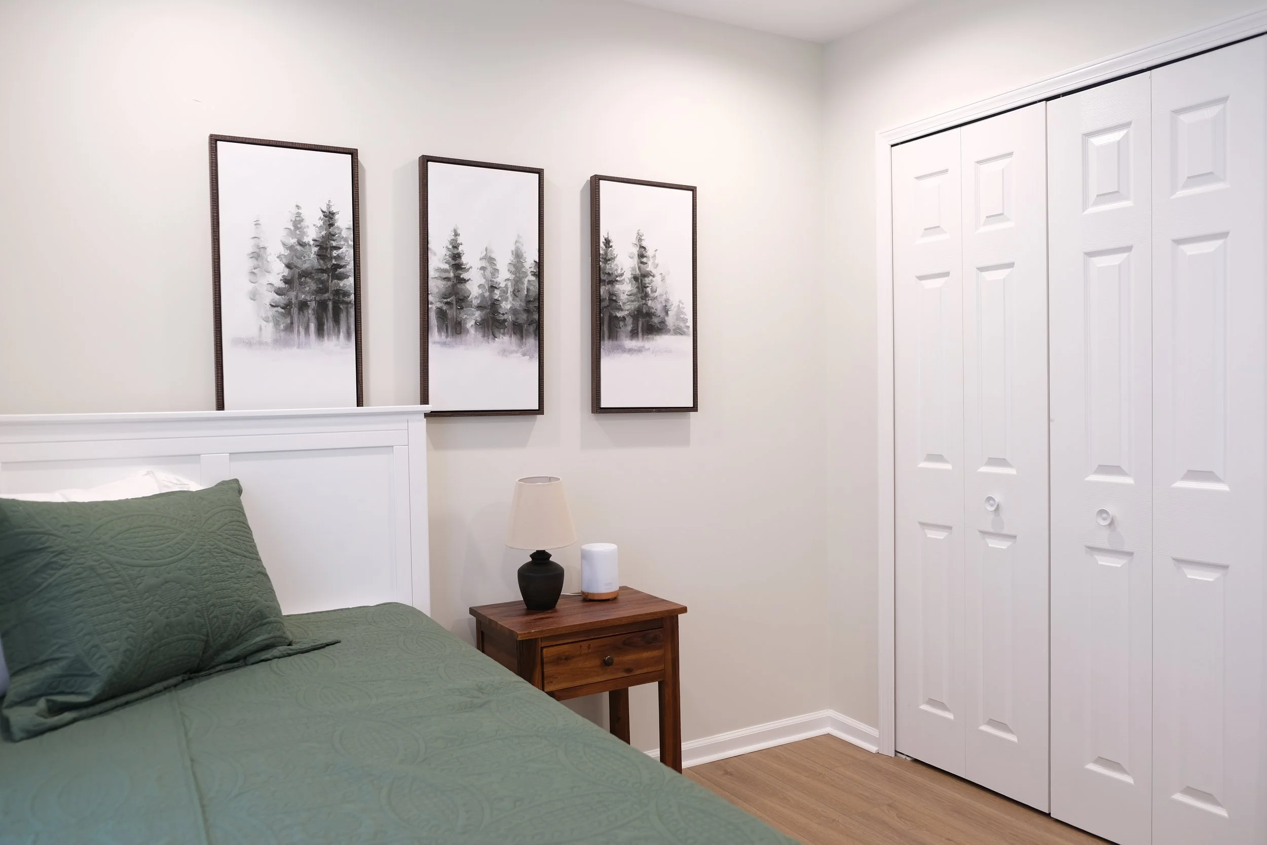 A bedroom with a white headboard, green bedspread and pillows, three framed black and white landscape art prints above the bed, a wooden nightstand with a lamp and a smart speaker, and white double closet doors.