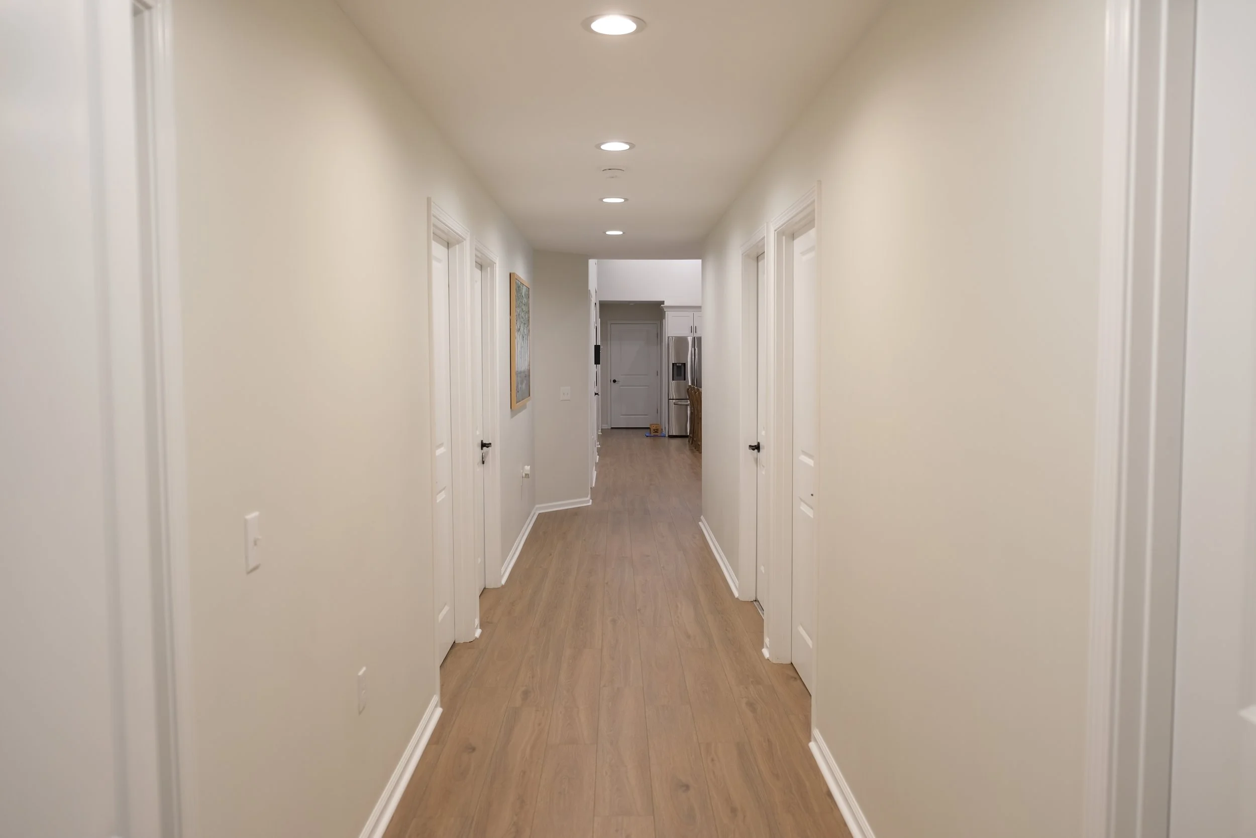 A hallway with beige walls, hardwood flooring, white doors on both sides, and recessed ceiling lights leading to a kitchen area with stainless steel appliances in the background.