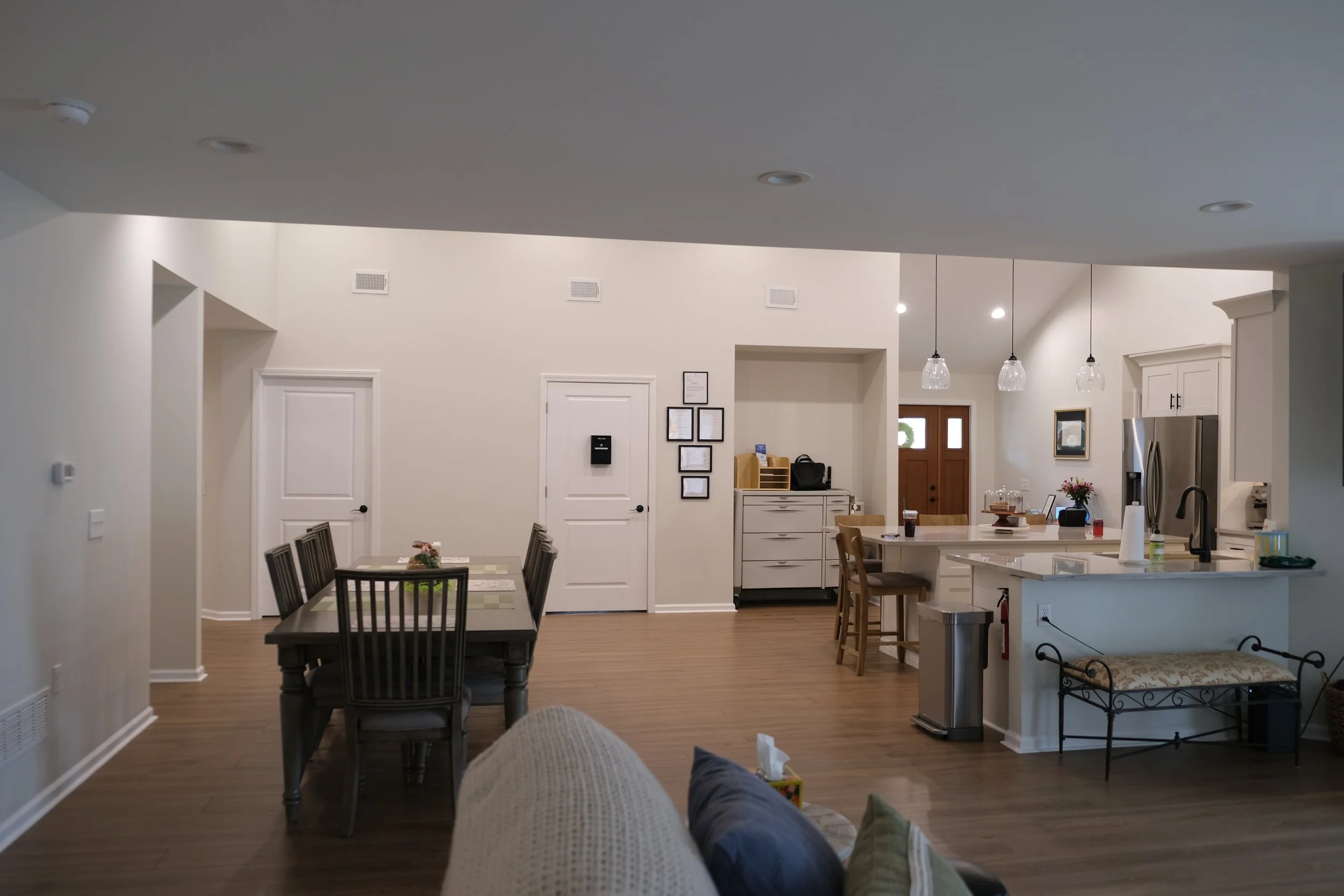 Open-concept living and dining area with white walls, hardwood floors, a dining table with six chairs, kitchen island with three bar stools, and pendant lights.