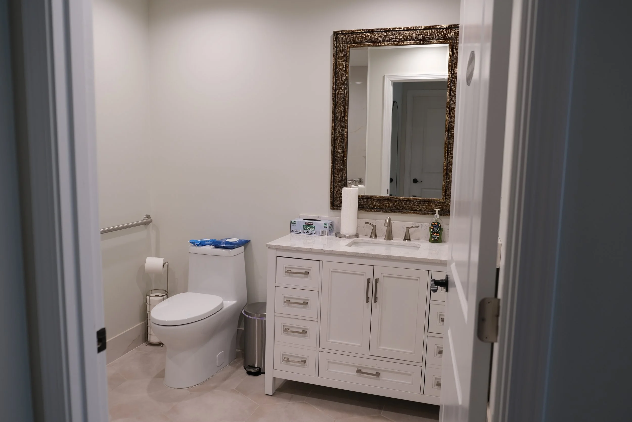 A bathroom with a toilet and a white vanity with a marble top, mirror, and various toiletries on the countertop.