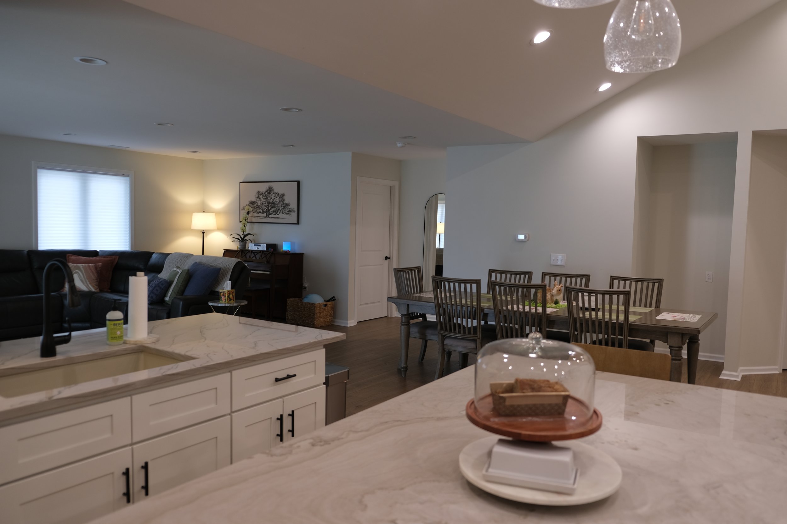 Open-concept kitchen and living room with white cabinetry, marble countertops, dining table, and a seating area with a couch, lamp, and wall art.