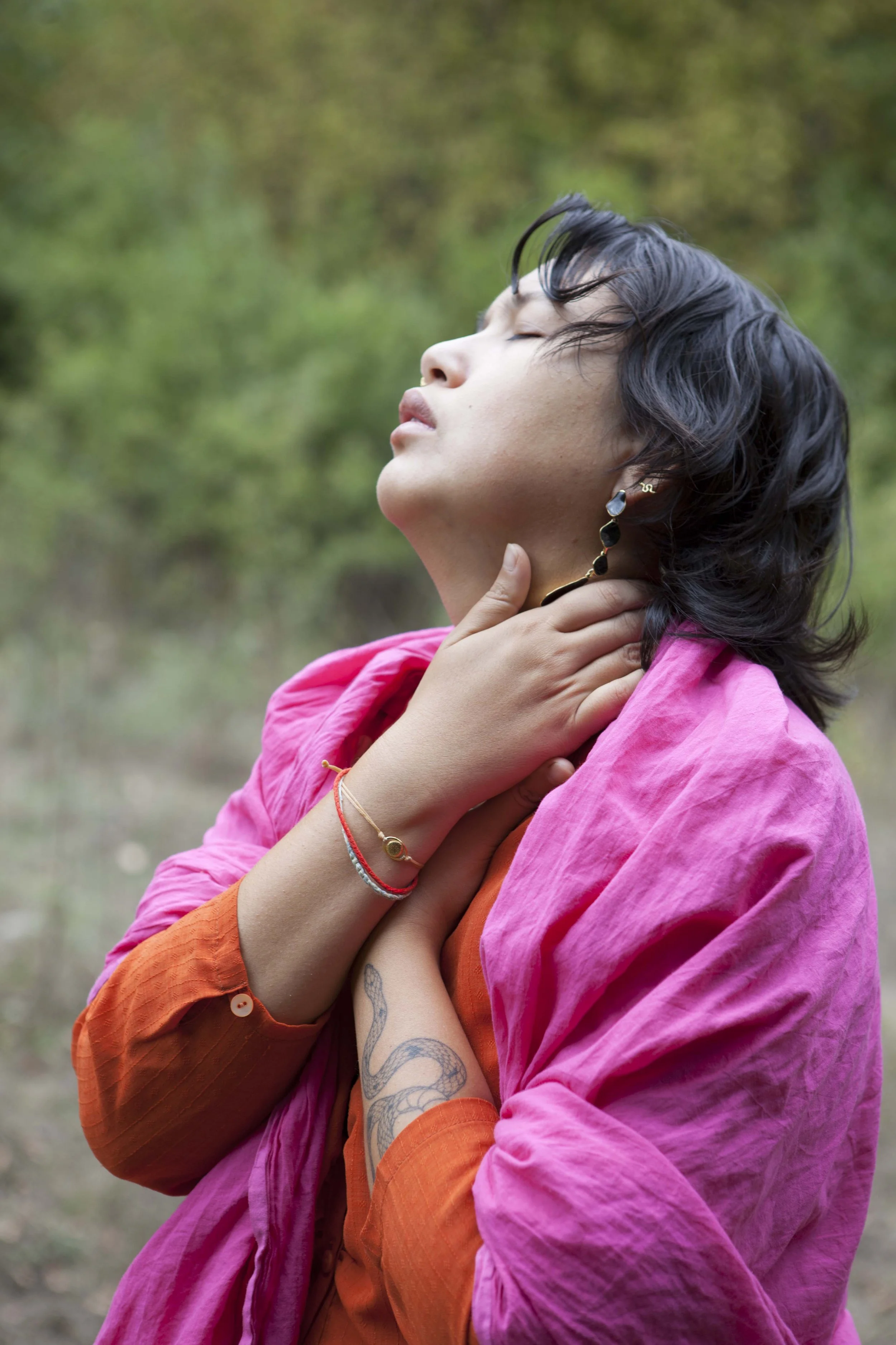 A woman with short black hair, wearing a pink shawl and an orange top, is outdoors with her eyes closed and her hand on her neck as if experiencing emotion or relief, with a green, blurred background.