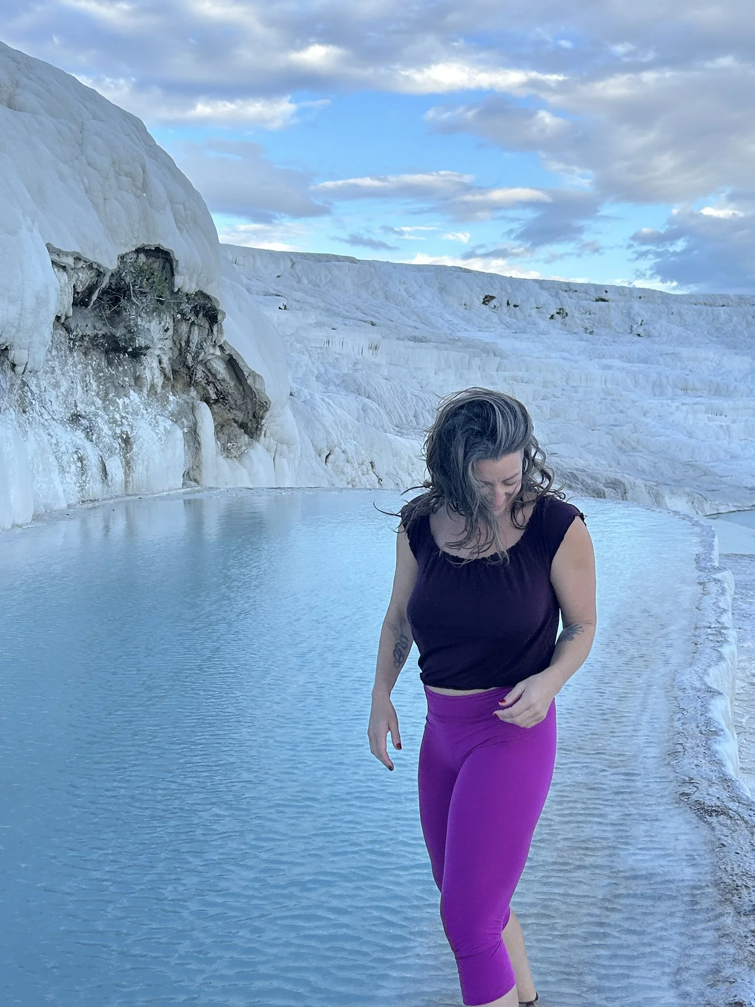 Woman with curly hair wearing a black top and bright pink leggings walking near the edge of a hot spring with white mineral deposits and rocky cliffs in the background.