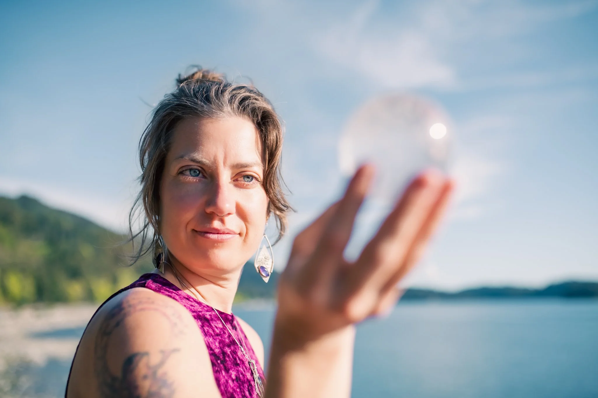 A woman with short brown hair and blue eyes holding a crystal ball outdoors near water, wearing a purple sleeveless top and earrings, with mountains and a blue sky in the background.