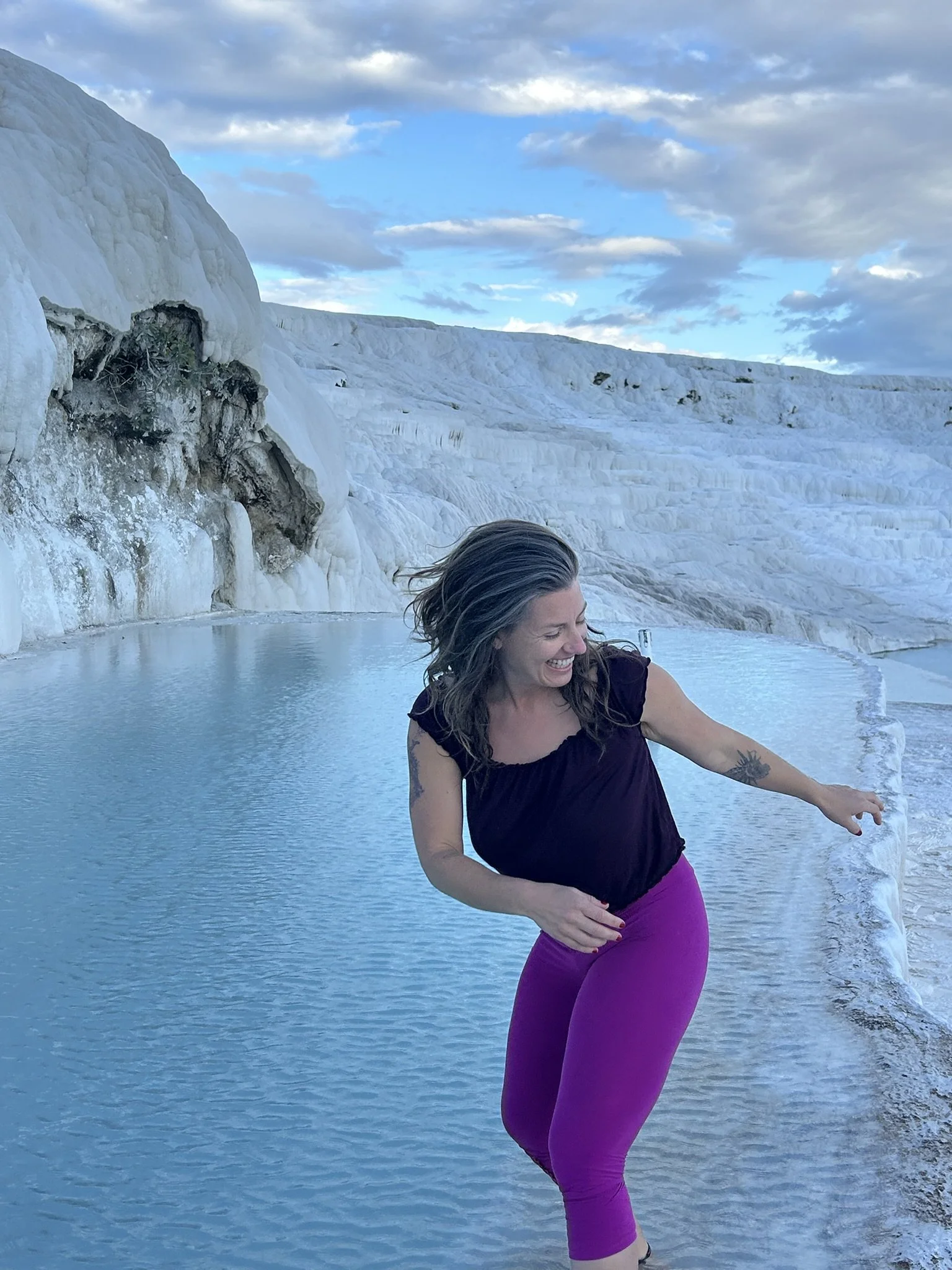 A woman with dark hair in a black top and bright purple leggings is smiling and playing in a hot spring surrounded by white mineral deposits, with a cloudy sky above.