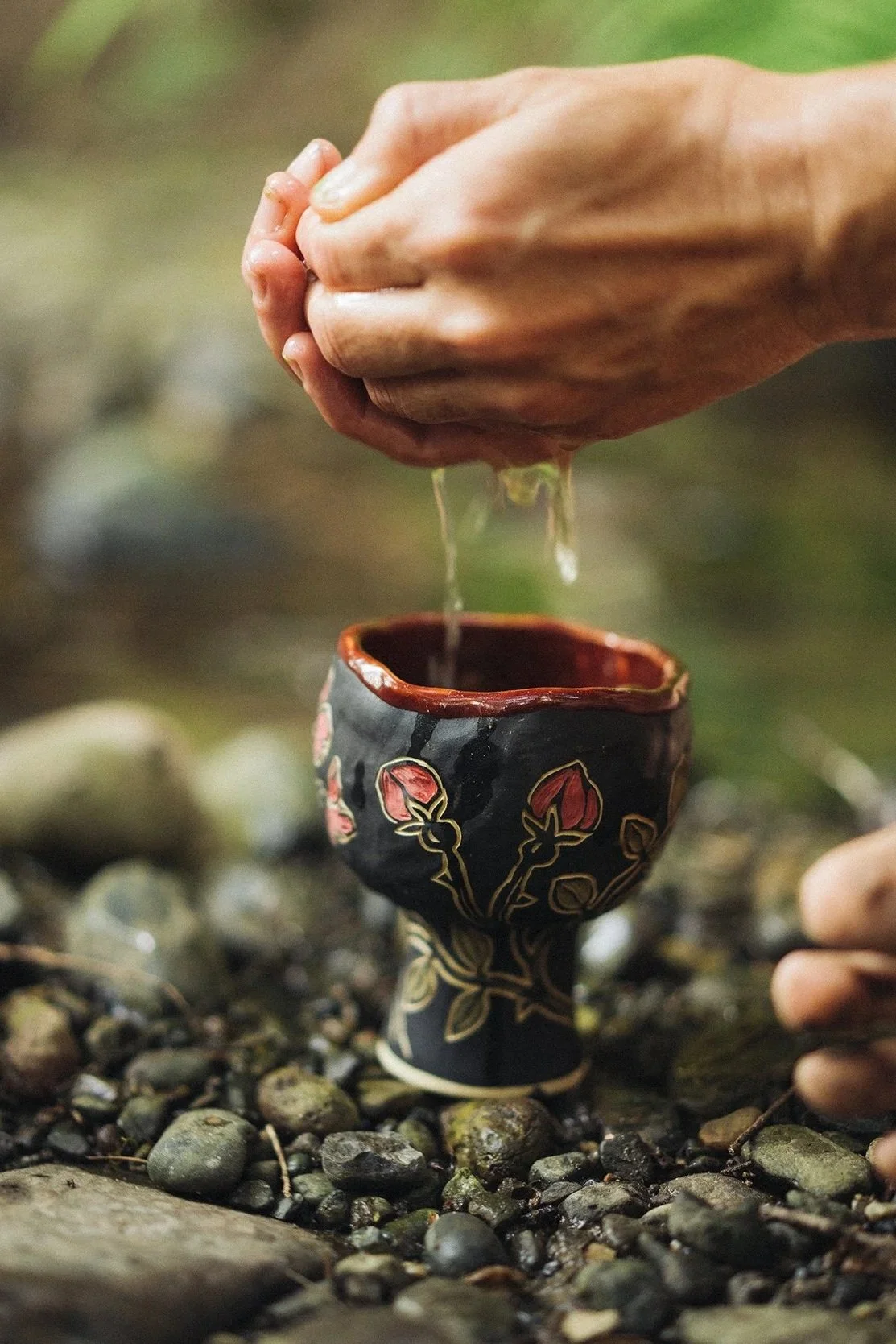 A person's hands holding water above a decorated ceramic cup with a floral pattern, outdoors on rocky ground.
