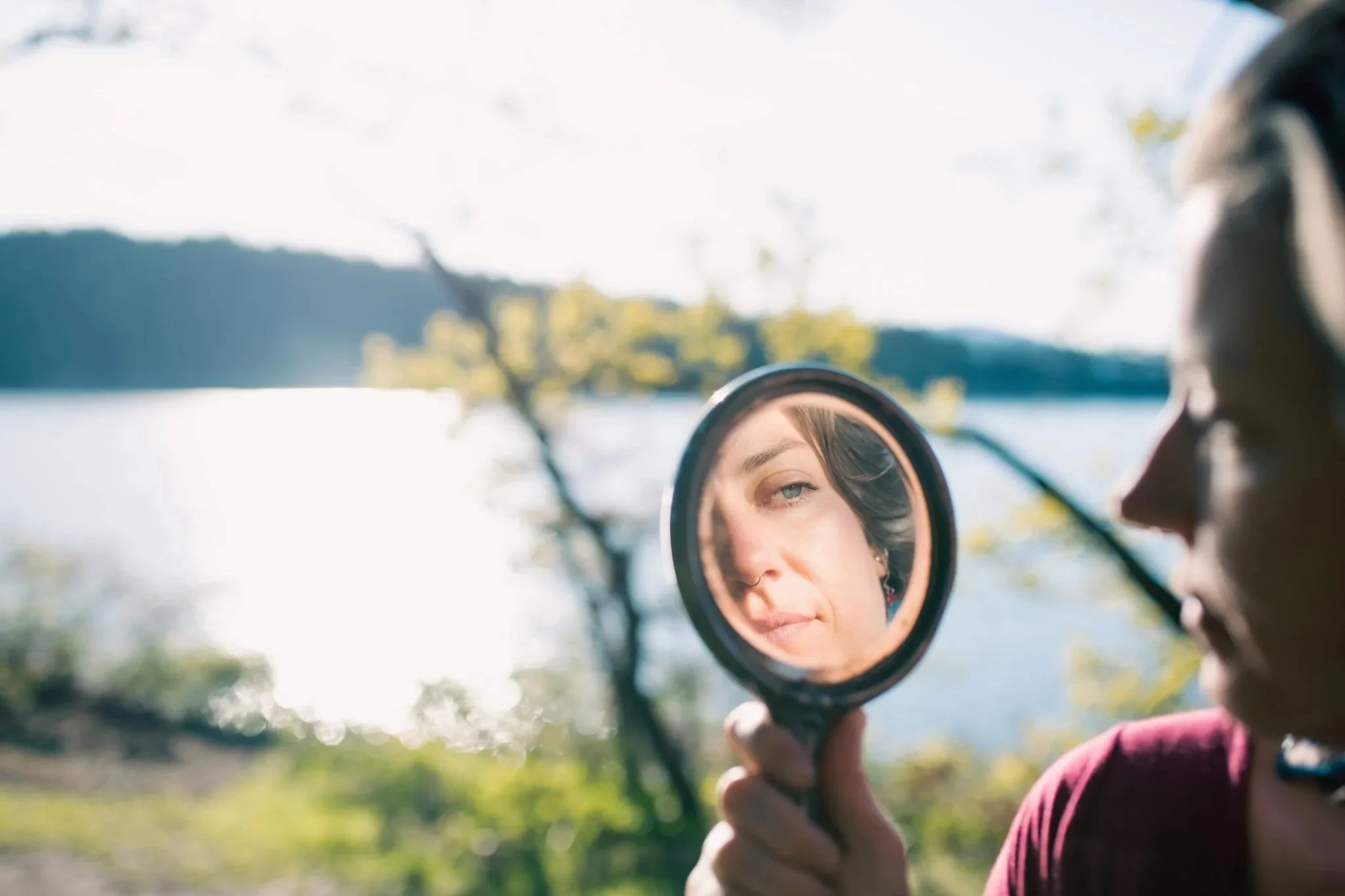 A person is holding a mirror and looking at their reflection with a lake, trees, and hills in the background.