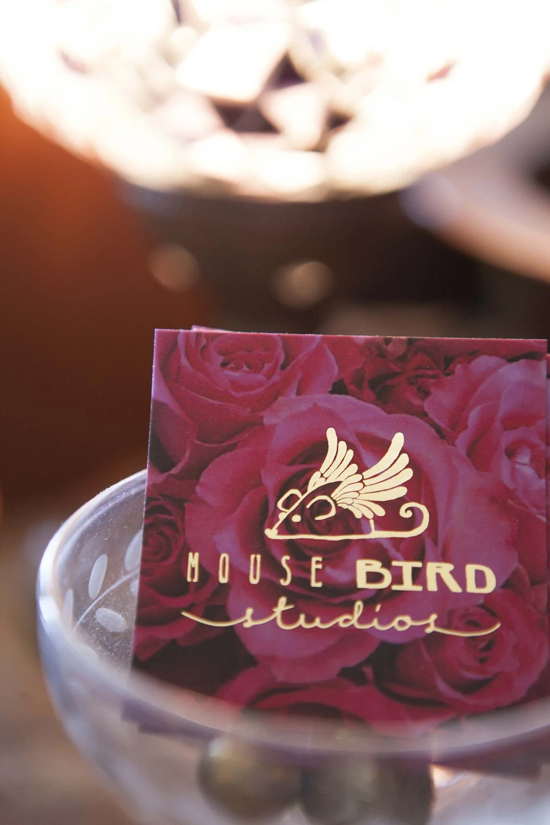 Business card for Mouse Bird Studios with a pink floral background, an outline of a bird with wings, and the studio's name in gold lettering inside a clear plastic container.