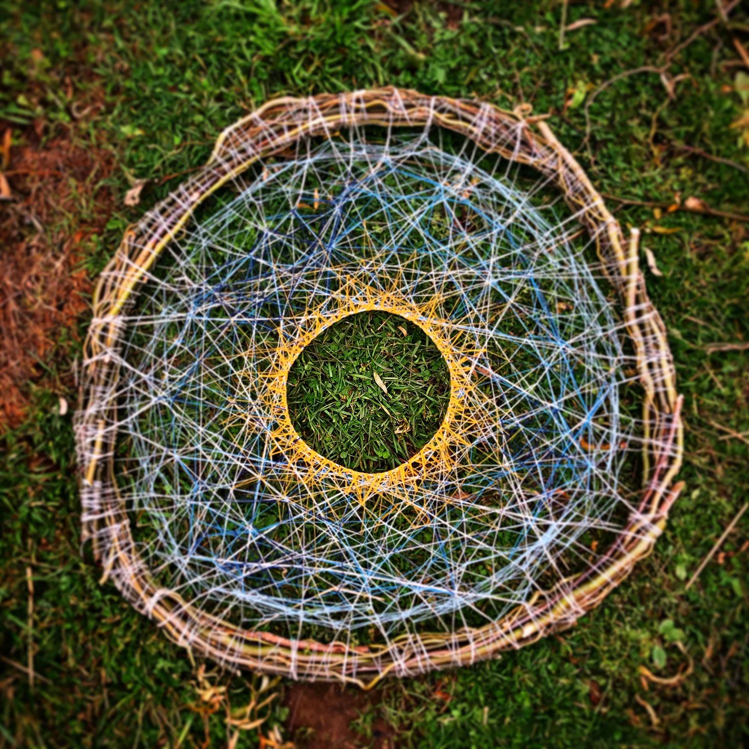 A woven dreamcatcher with intricate web patterns, lying on green grass.