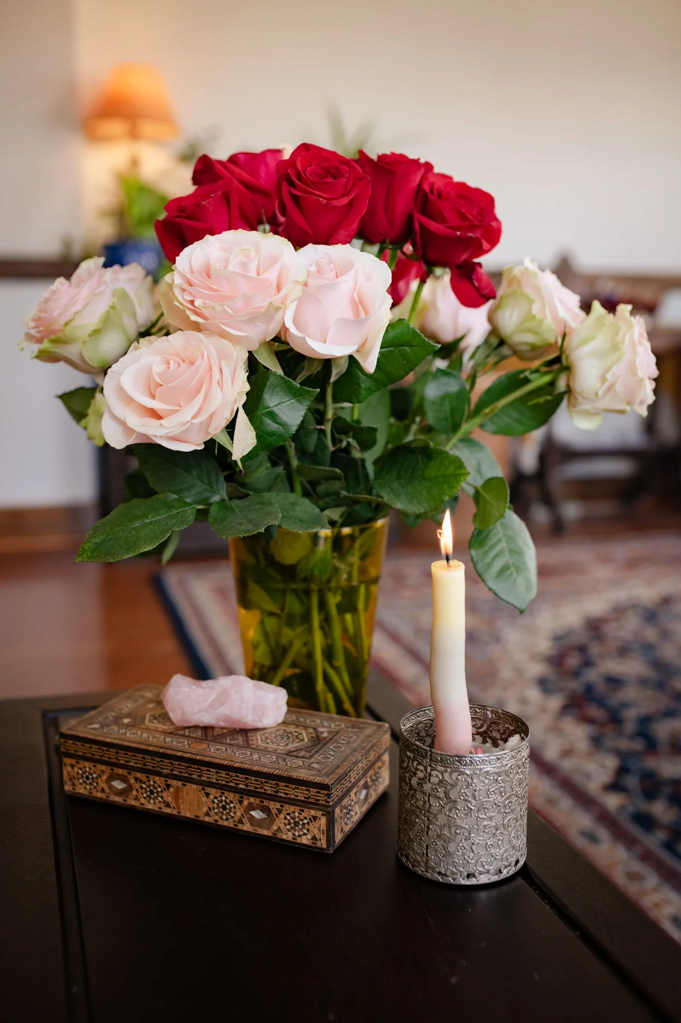 A bouquet of red, pink, and white roses in a glass vase on a table, with a lit candle in a decorative holder, a carved box with a pink crystal stone, in a living room with a rug and furniture in the background.
