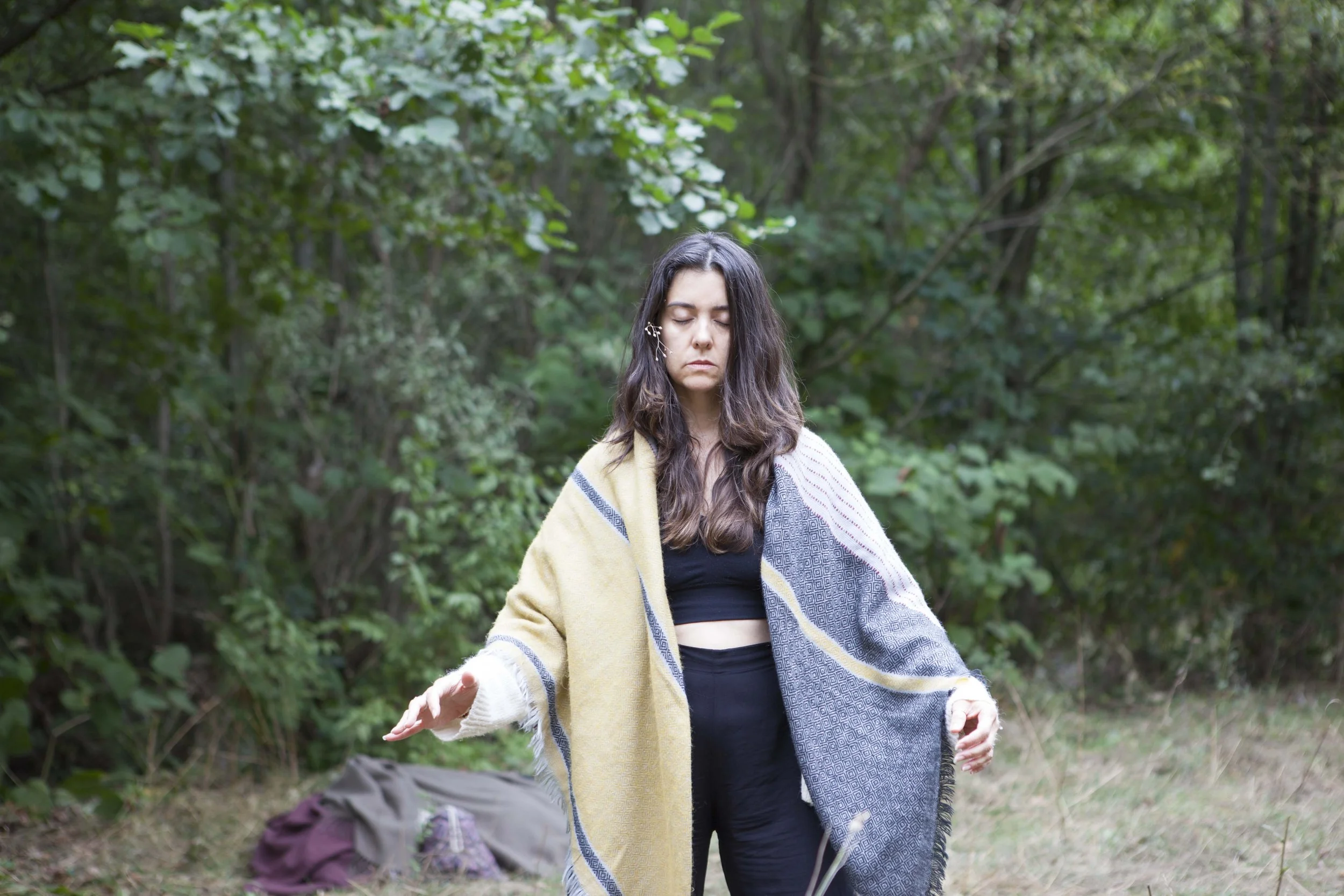 Young woman with long dark hair standing outdoors in a wooded area, eyes closed, with a large beige and gray blanket draped over her shoulders, and a backpack on the ground behind her.