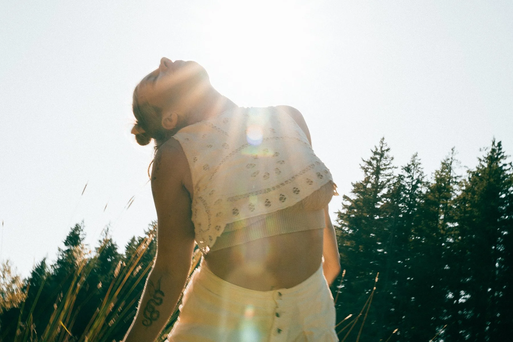 A woman outdoors with the sun behind her, wearing a light-colored sleeveless top and high-waisted pants, with trees visible in the background.