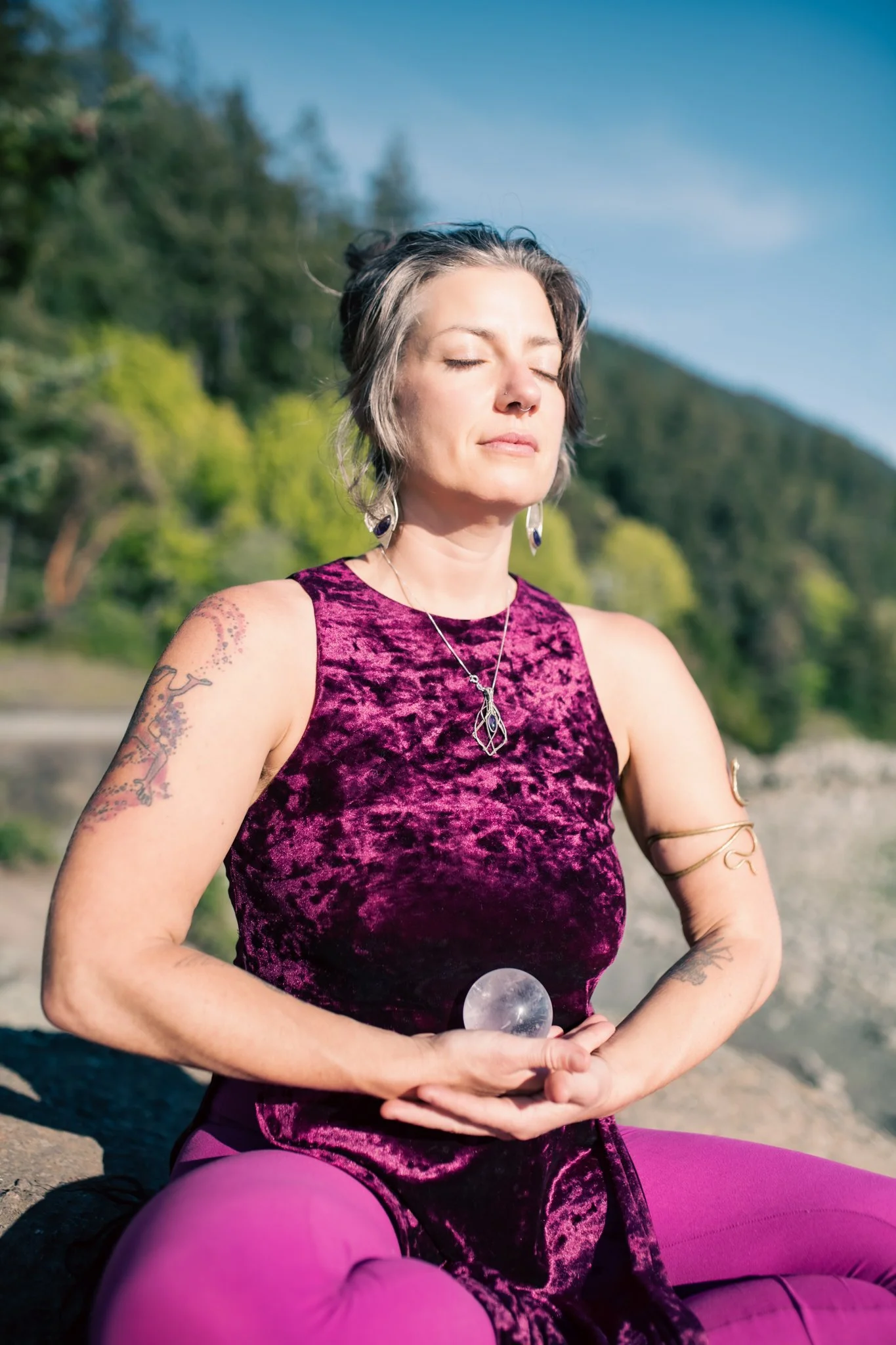 A woman with short gray hair, tattoos, and jewelry practices yoga outdoors on a sunny day, holding a clear ball in her hands with closed eyes.