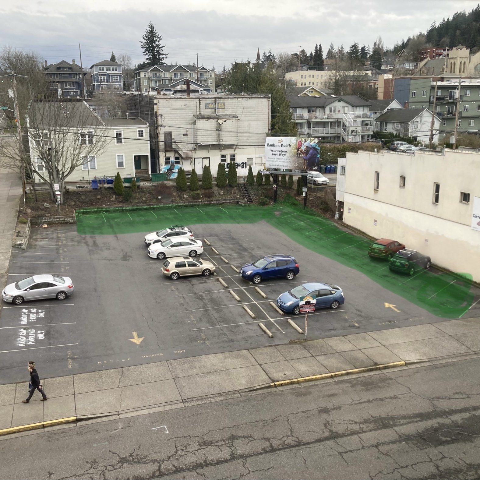 Parking lot with several parked cars and a person walking on sidewalk, with buildings and trees in the background.