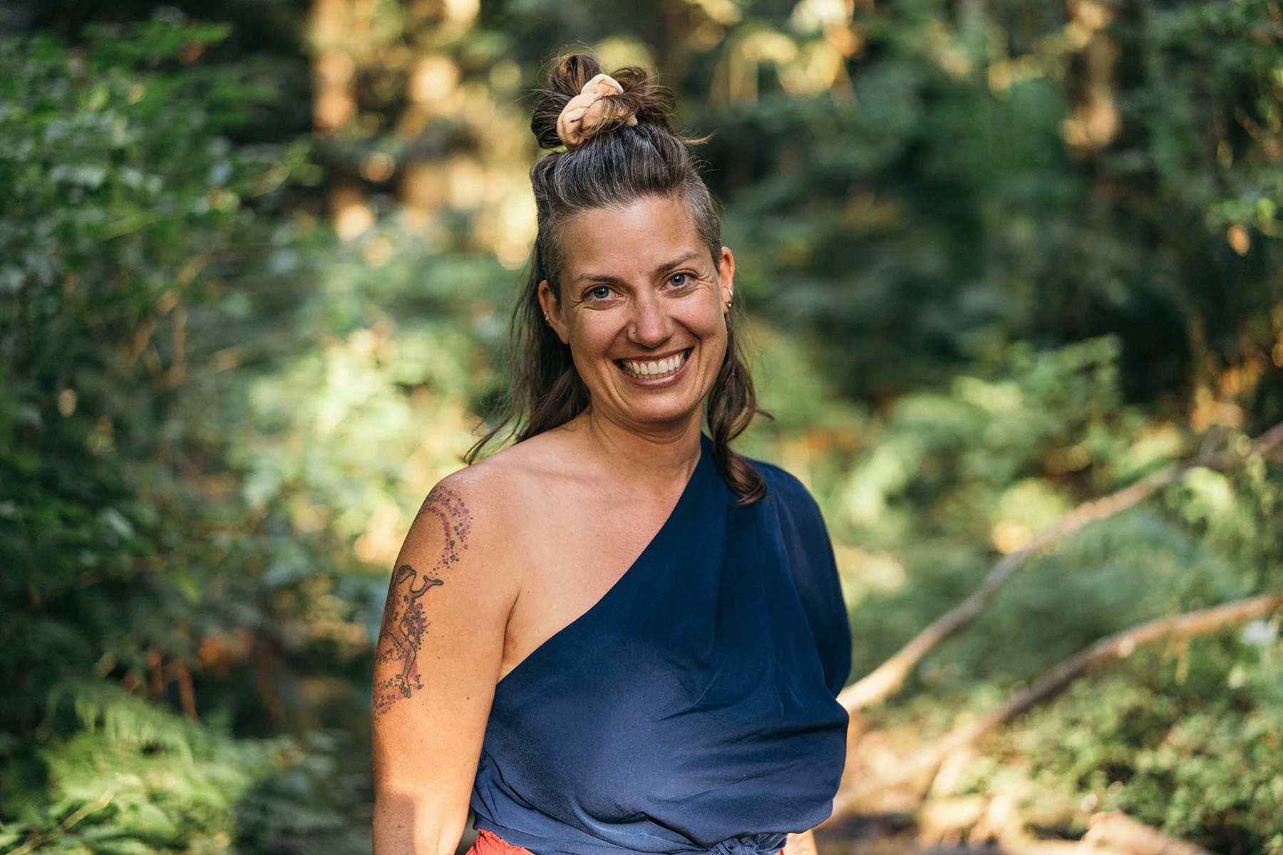 A smiling woman in a blue one-shoulder top standing outdoors in a wooded area with green trees in the background.