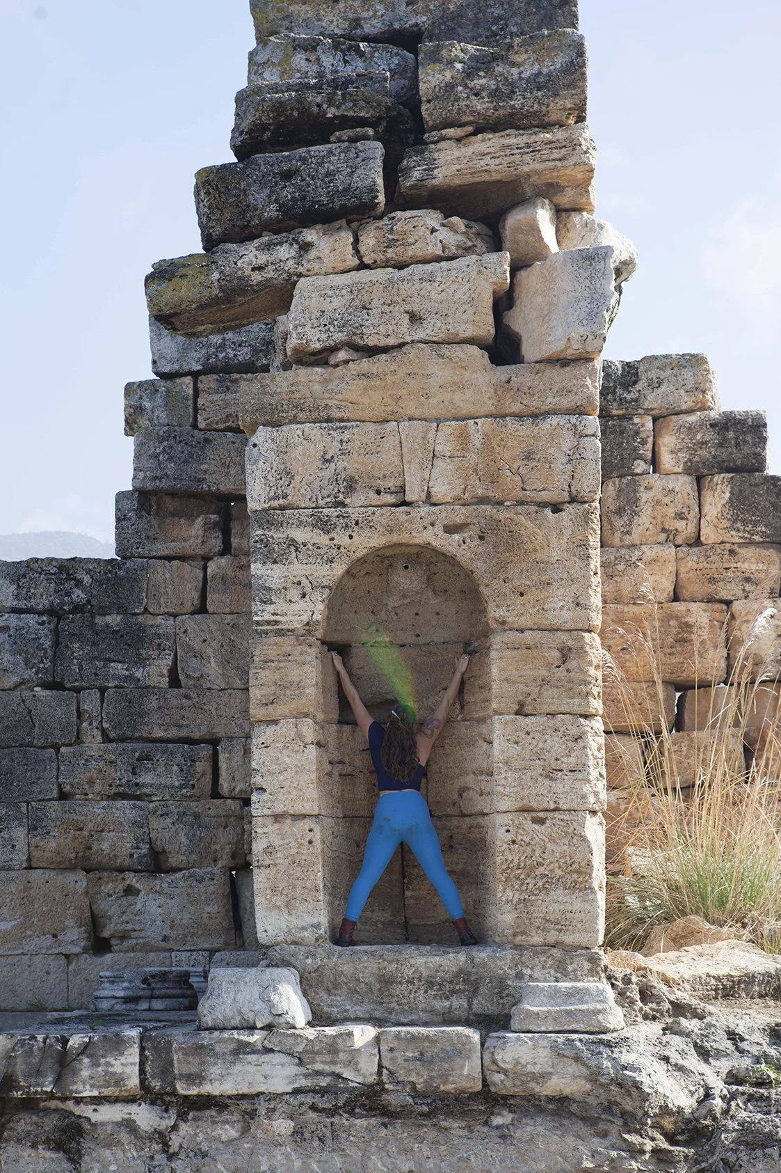 A woman in blue pants and a dark shirt is climbing or posing against an ancient stone wall with a small arched alcove, outdoors near a grassy area.
