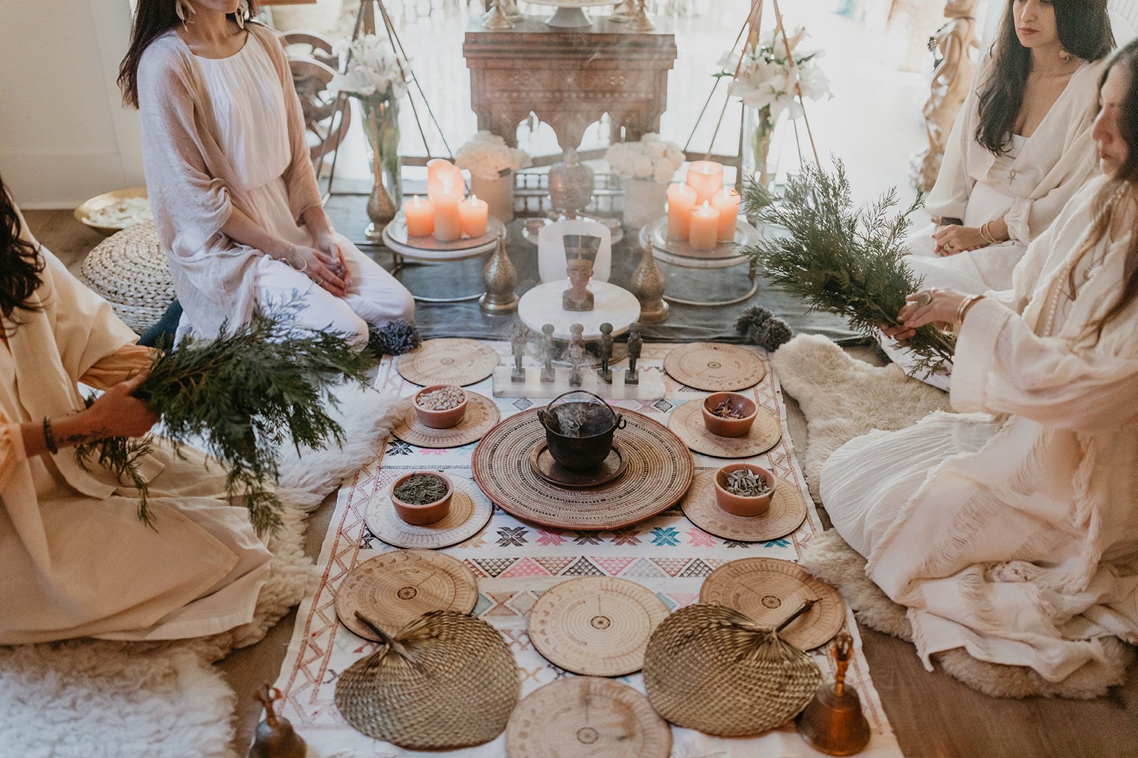 Women sitting on the floor in a circle during a spiritual or meditation session, with candles, plants, and mystical objects on a low table and a patterned rug.