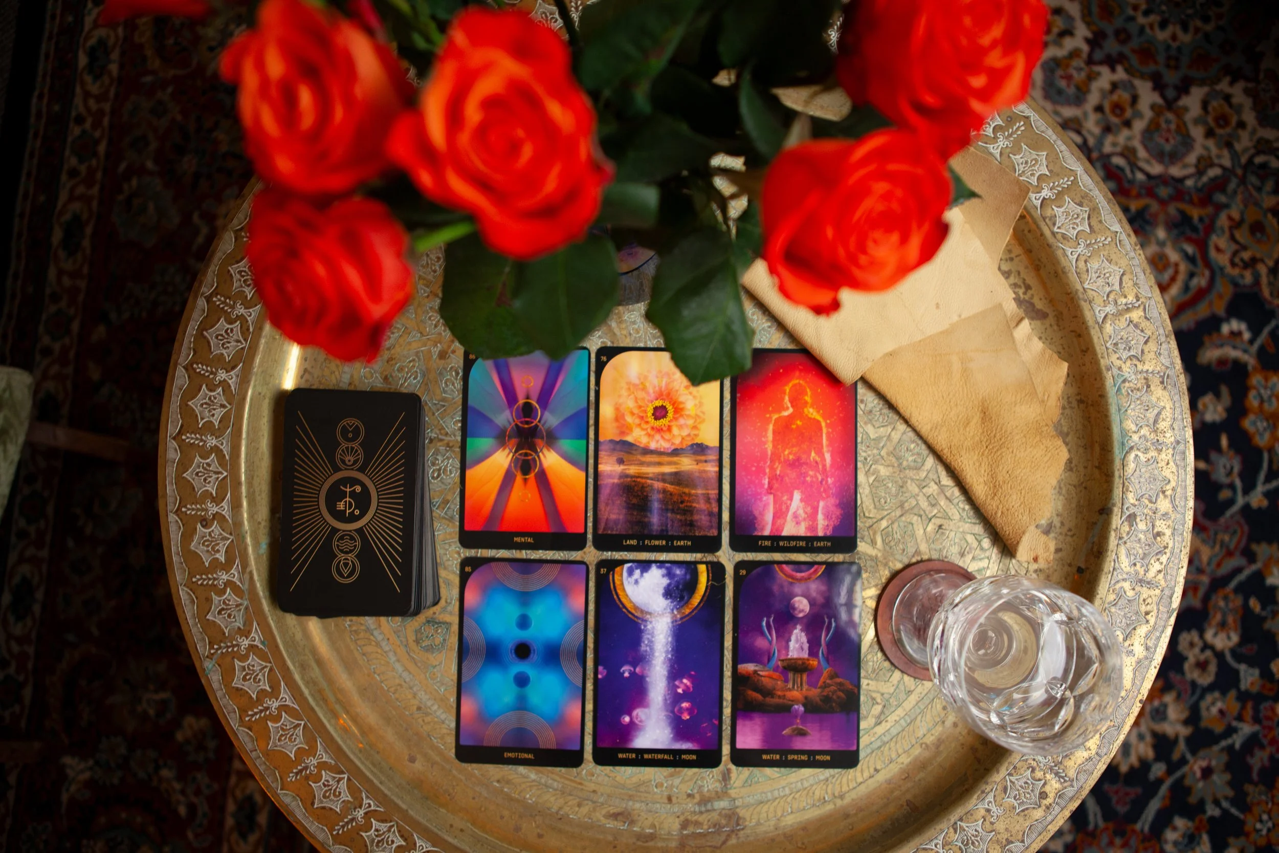 A tarot card spread on a round table with a bouquet of orange roses, a glass of water, and a beige cloth. The spread includes six tarot cards with vibrant colors and mystical imagery.