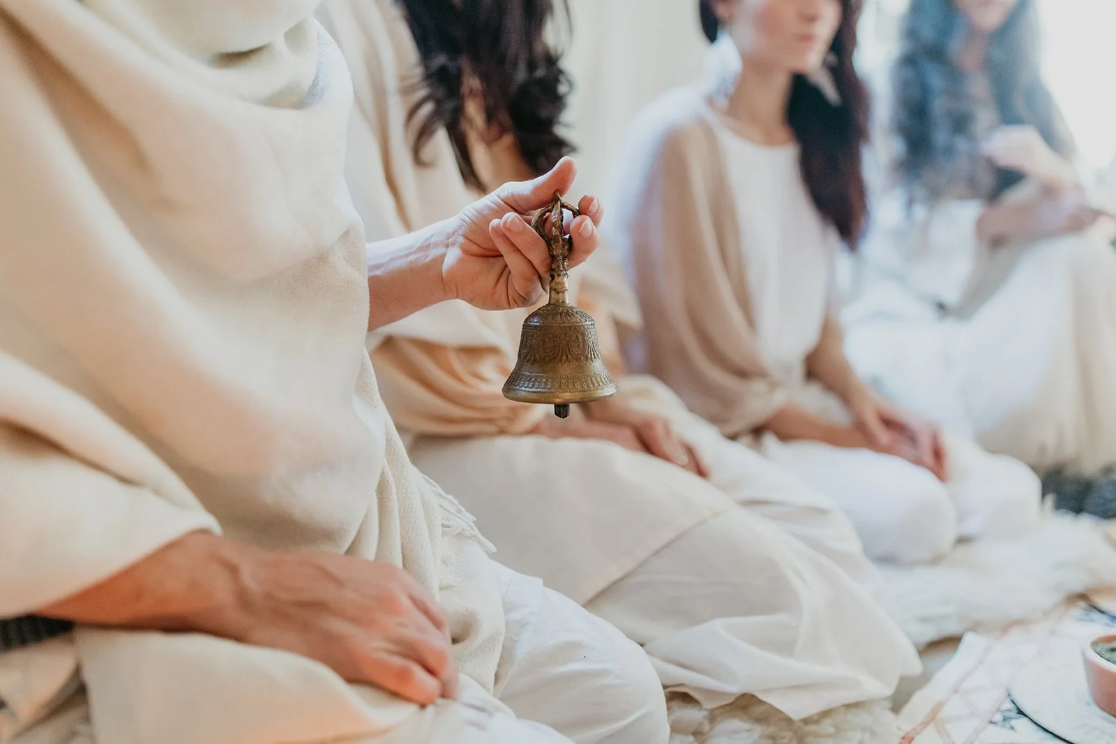 A woman holding a brass bell while sitting with others dressed in white, engaged in a group activity.
