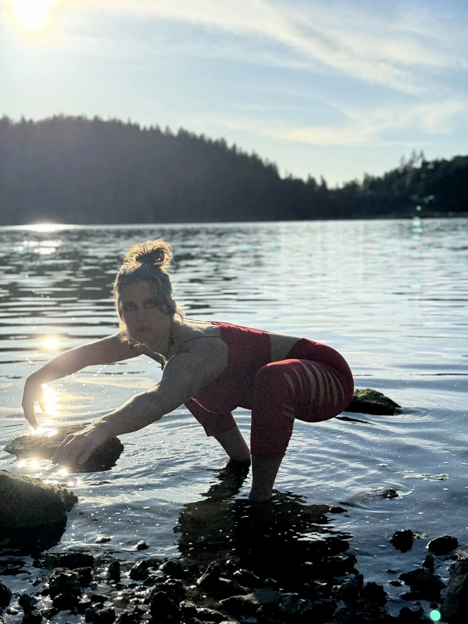 A woman in red workout clothes crouching in shallow water near rocks with a scenic lake and hills in the background, during sunset or sunrise.