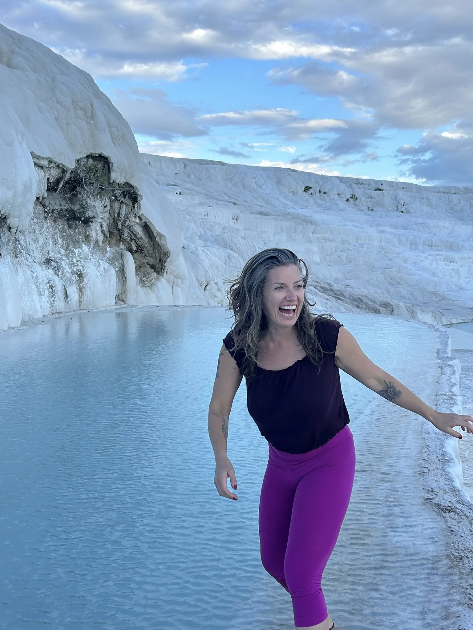 A woman with brown hair, tattoos, and wearing a black top and purple pants, appears to be laughing or expressing joy while standing near a pool of water at Pamukkale in Turkey, with white terraces and blue sky in the background.
