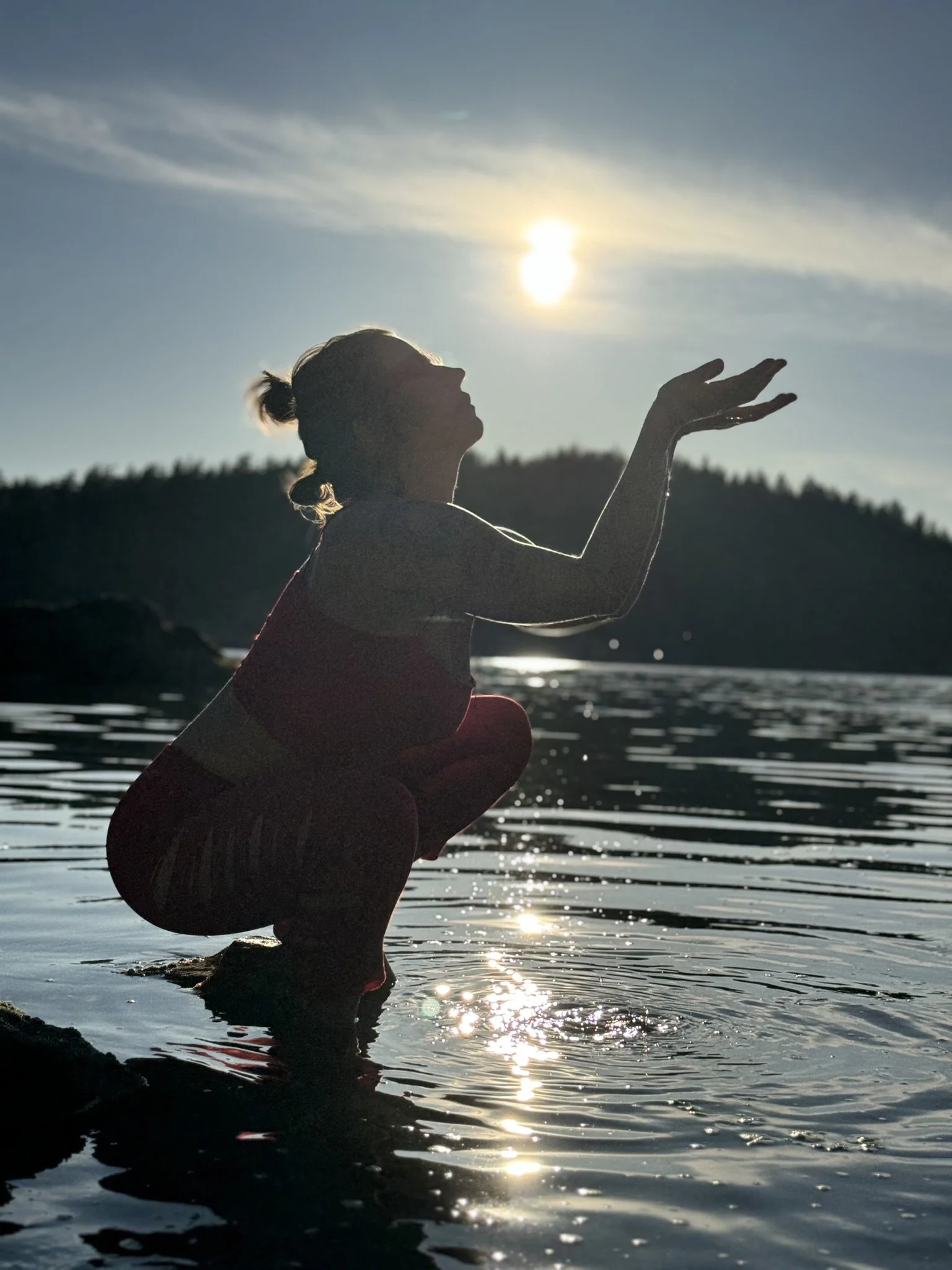 A person crouching in water at sunset, with hills in the background, silhouetted against the sky and sun.
