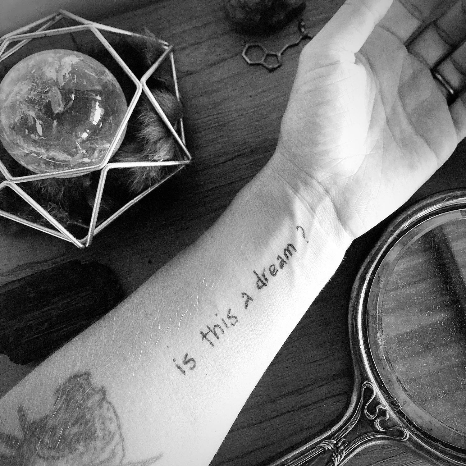 A person's arm with the text 'is this a dream?' tattooed on it. On the table are a geometric glass with a moon-like object inside, scissors, and a decorative mirror.