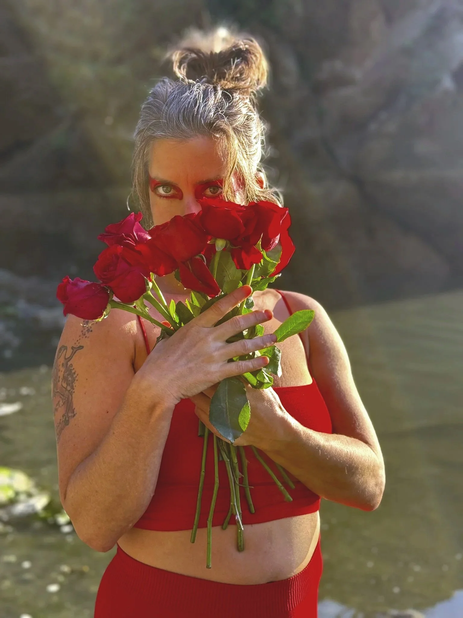 woman with a bun hairstyle holding a bouquet of red roses, wearing a red top, with a rocky outdoor background.