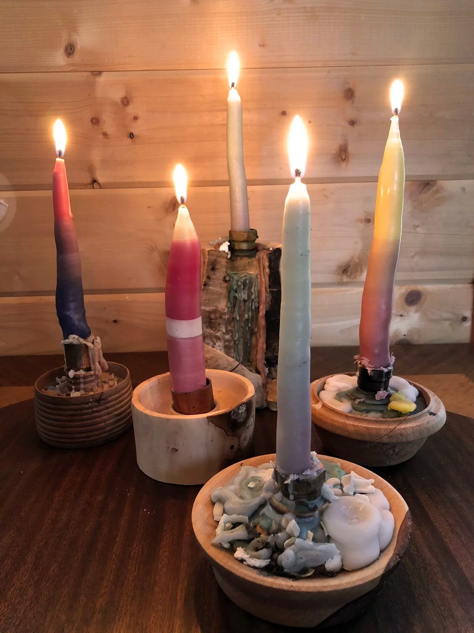Five colorful candles on wooden and ceramic holders, some lit with flames, with a wooden wall background.