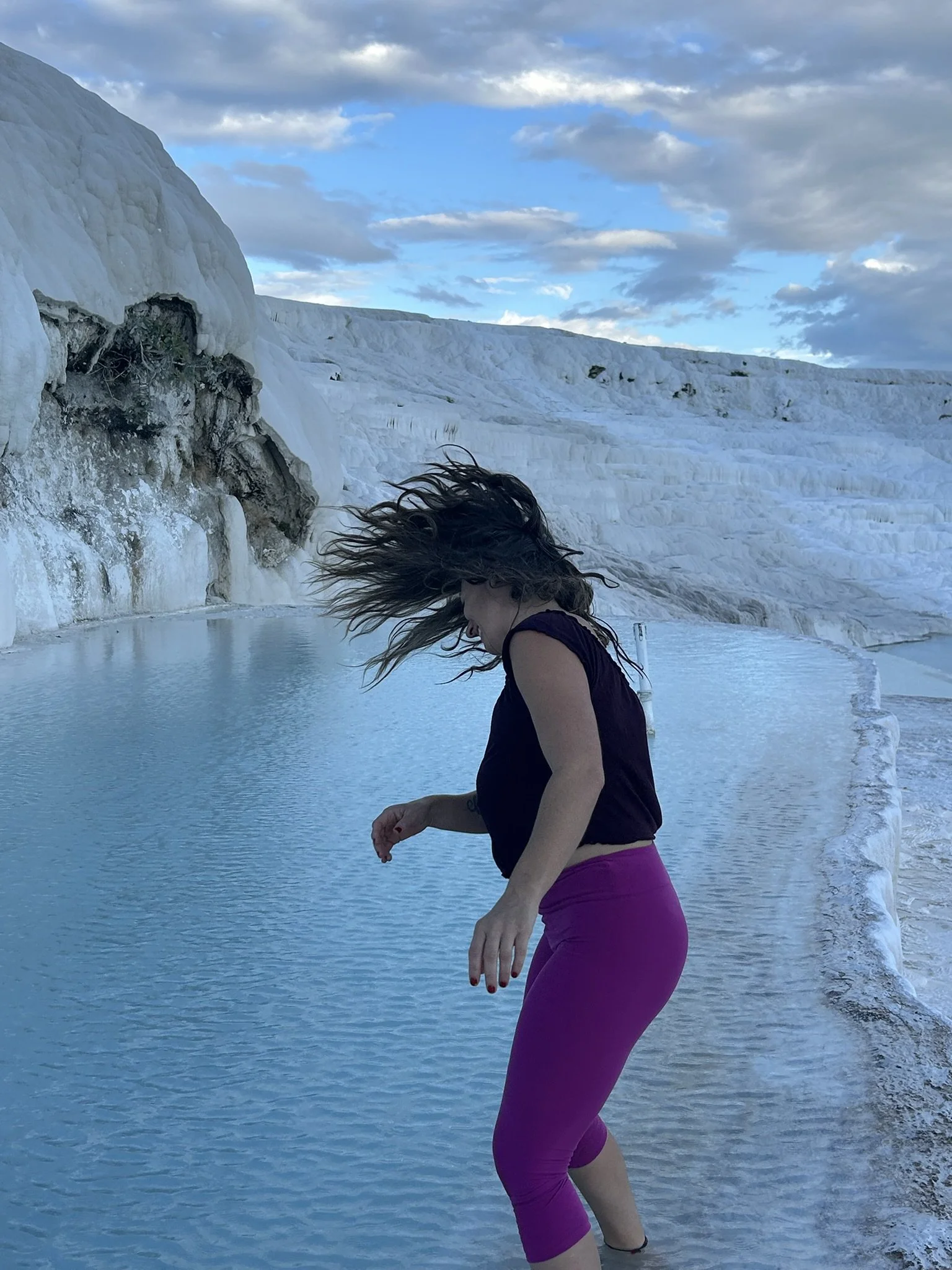 A woman with long hair, wearing a black top and purple leggings, standing in shallow water near a white, rocky cliff with a blue sky and clouds in the background.