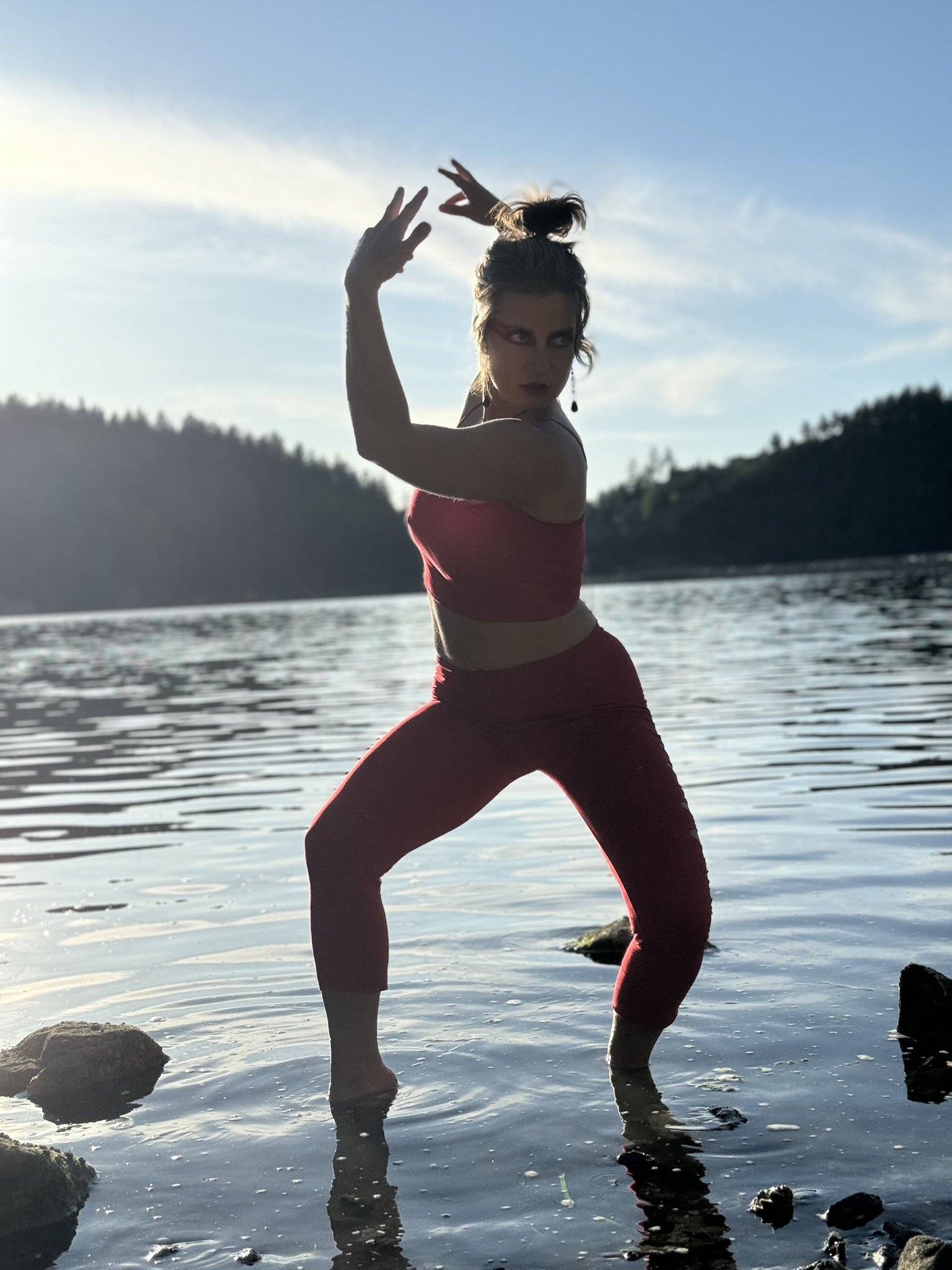 A woman in red clothing striking a dance pose in shallow water with mountains and a cloudy sky in the background.
