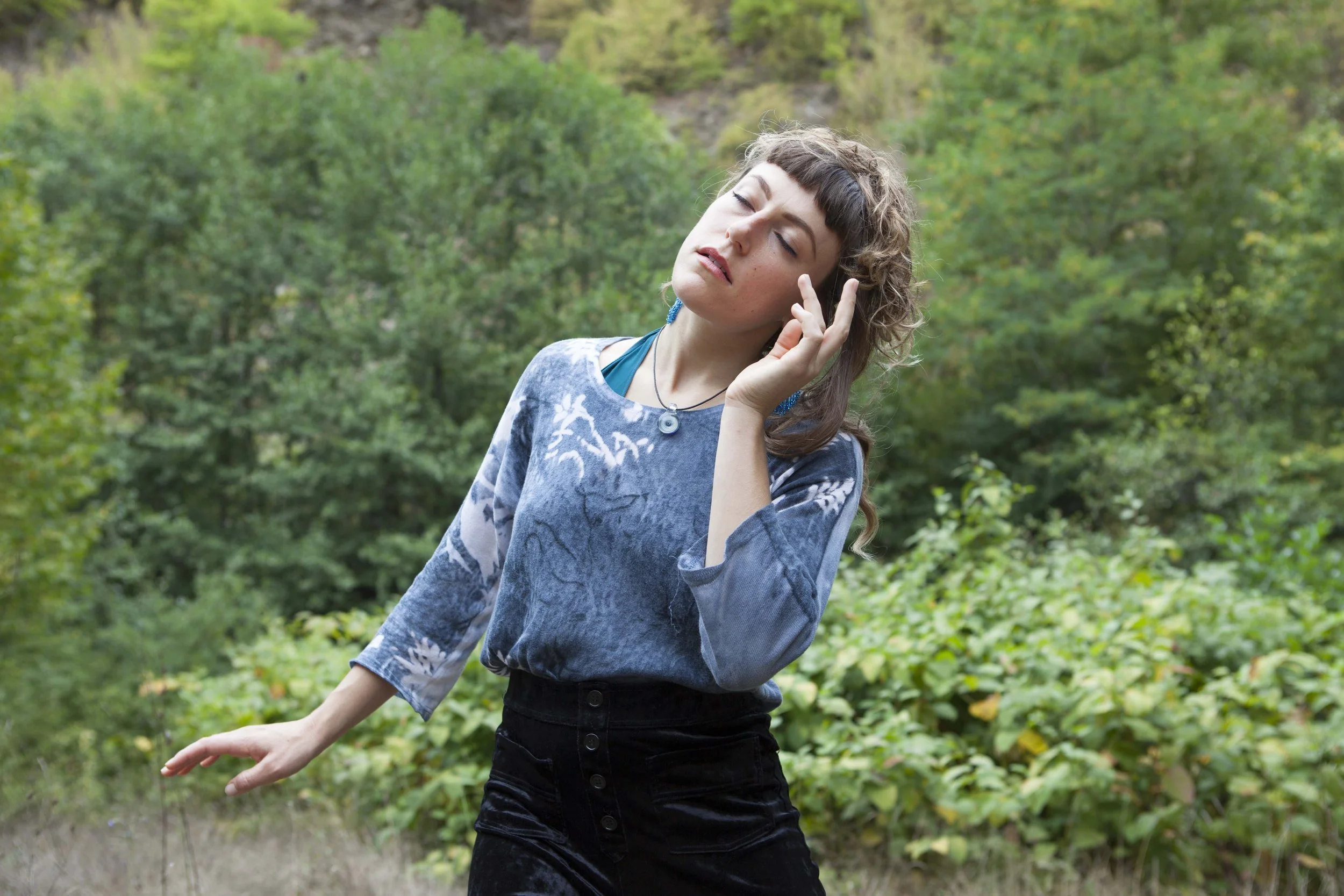 A woman with curly hair wearing a patterned blue top and black pants, standing outdoors surrounded by greenery, with her eyes closed and one hand touching her face.