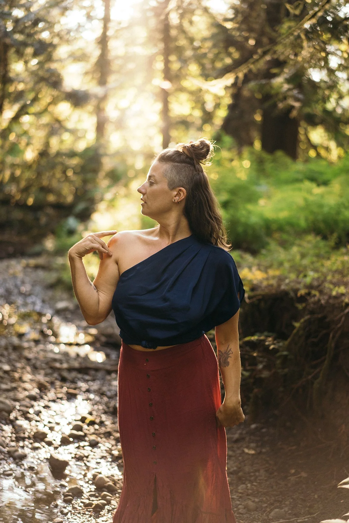 A woman standing outdoors in a forest during sunlight, wearing a navy blue top draped over one shoulder and a long red skirt with buttons, with her hair styled half-up and a tattoo on her arm.