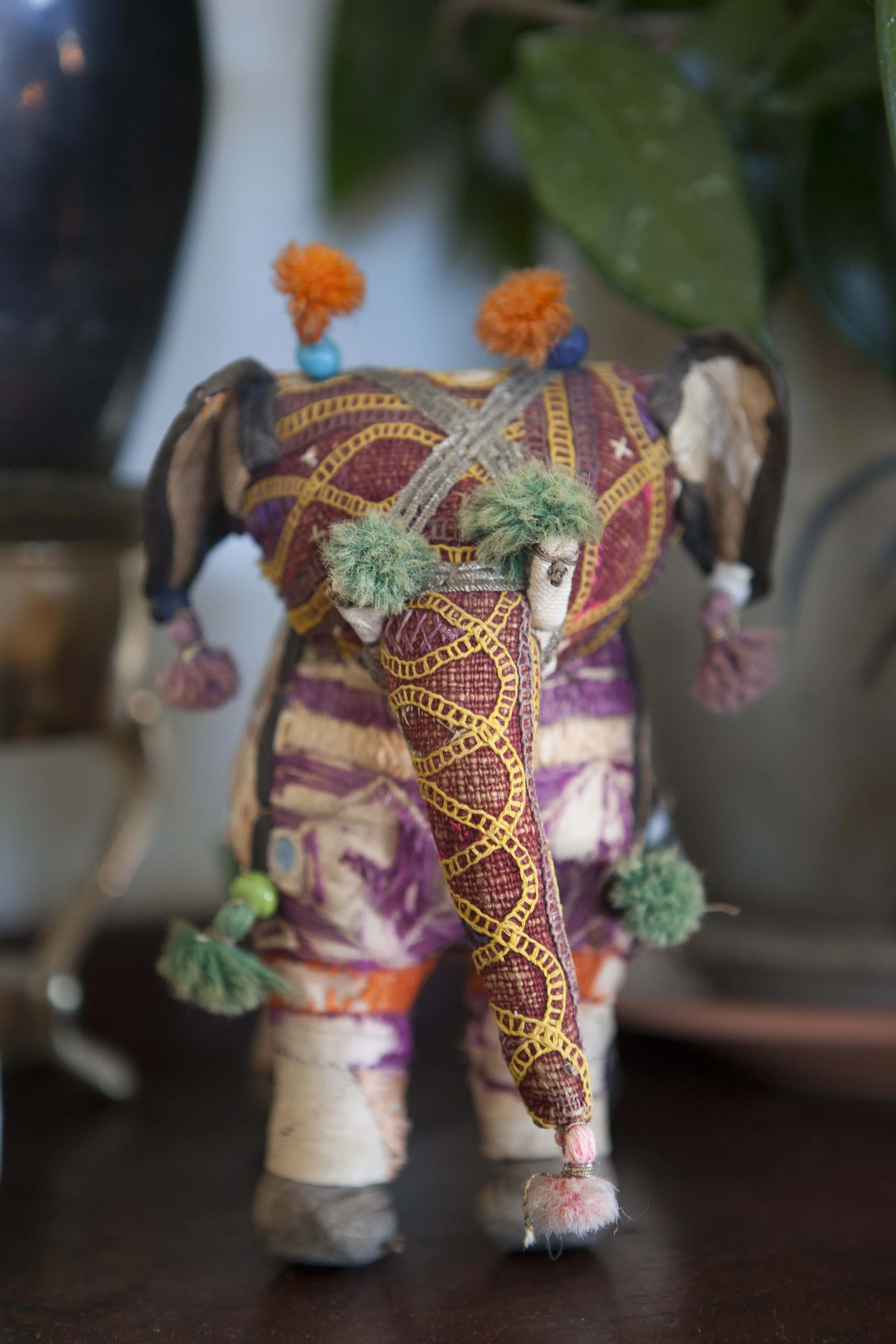 Decorative stuffed elephant with colorful fabric and detailed embroidery, adorned with colorful pom-poms and yarn decorations, standing on a wooden surface with plants in the background.
