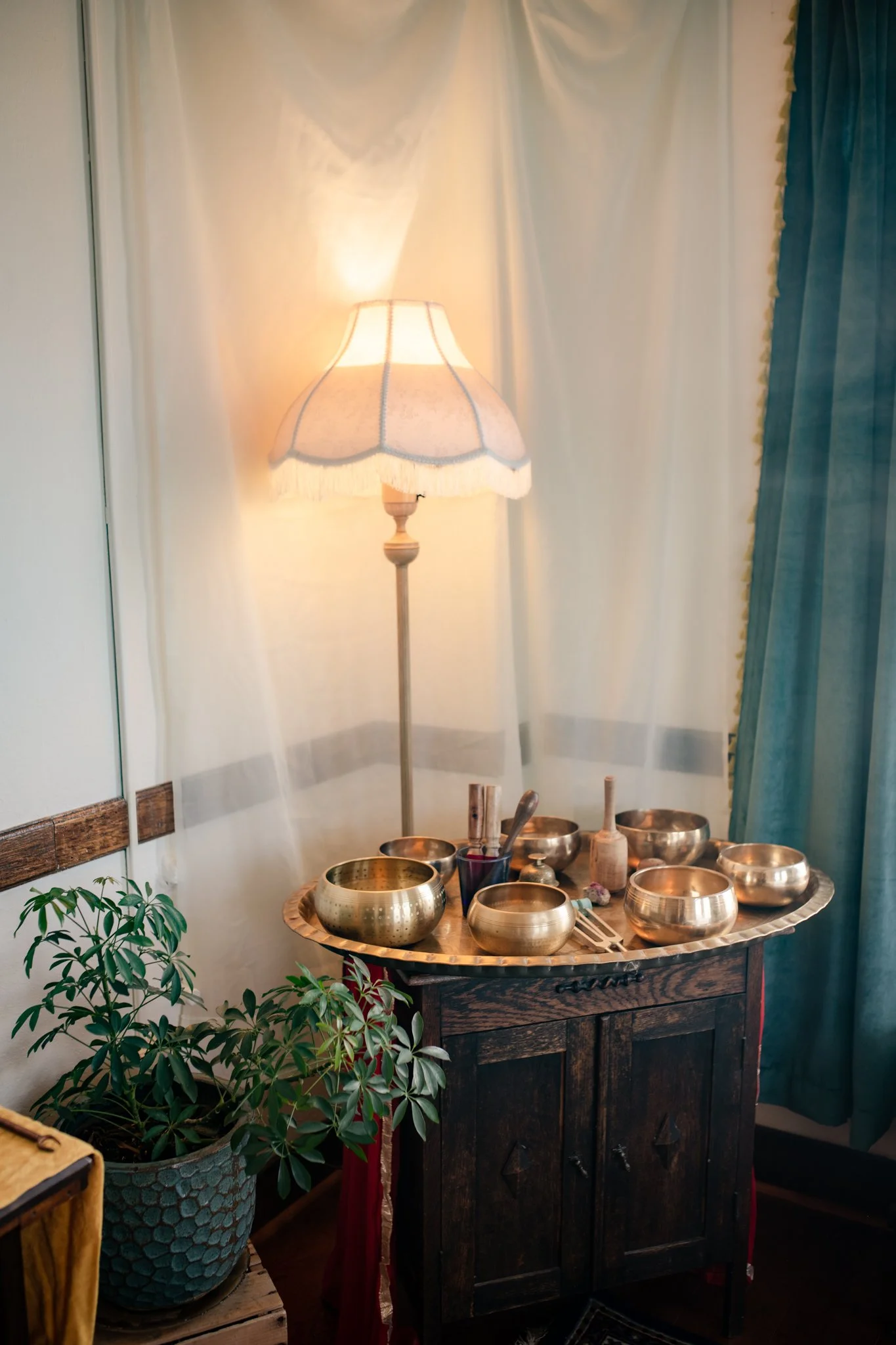 Vintage floor lamp illuminating a wooden table with seven brass singing bowls, hammer, and mallets in a cozy room with plants, curtains, and white drapes.