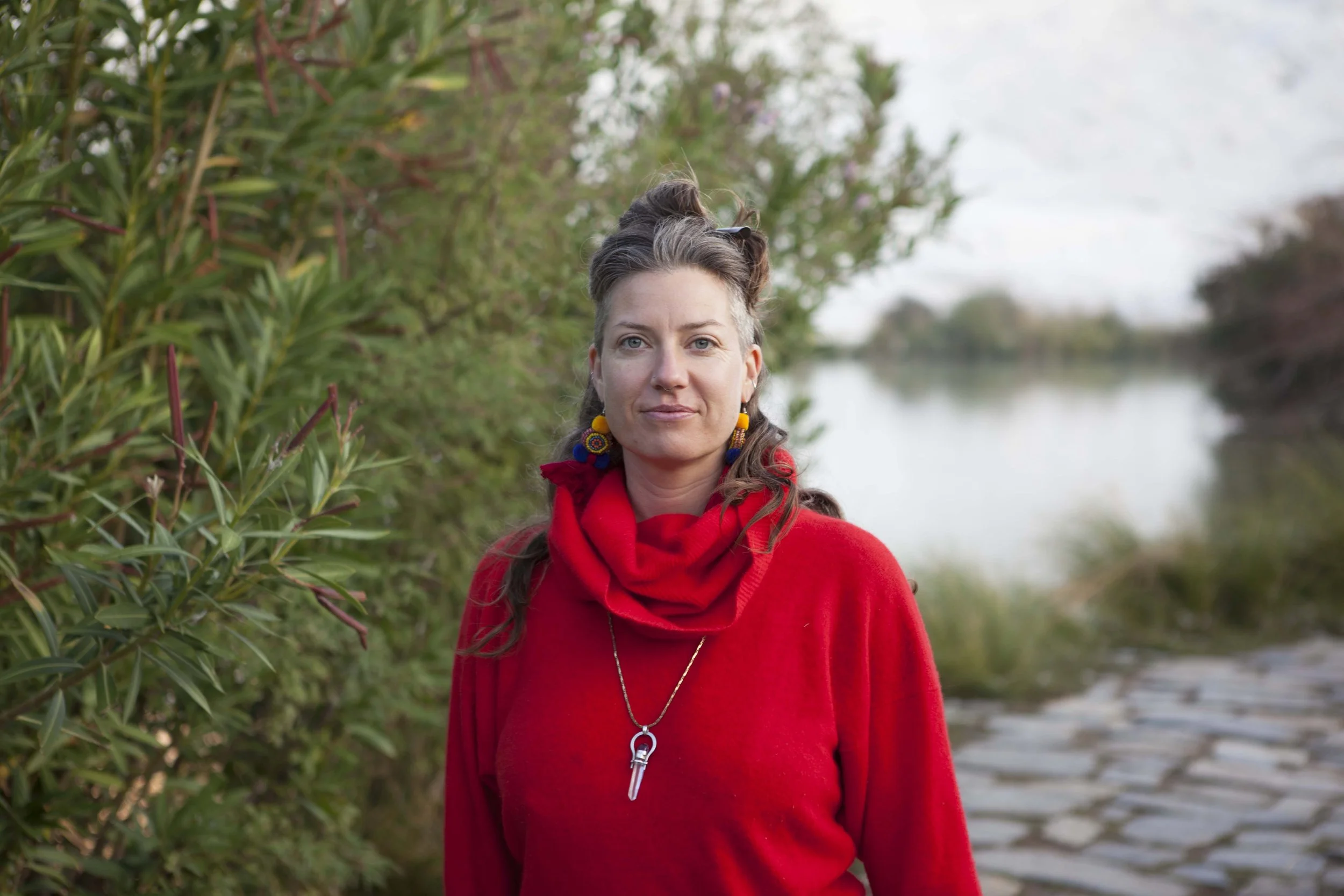 A woman with wavy brown hair in a castle trail, wearing a red sweater and necklace, standing near a body of water with trees and greenery in the background.