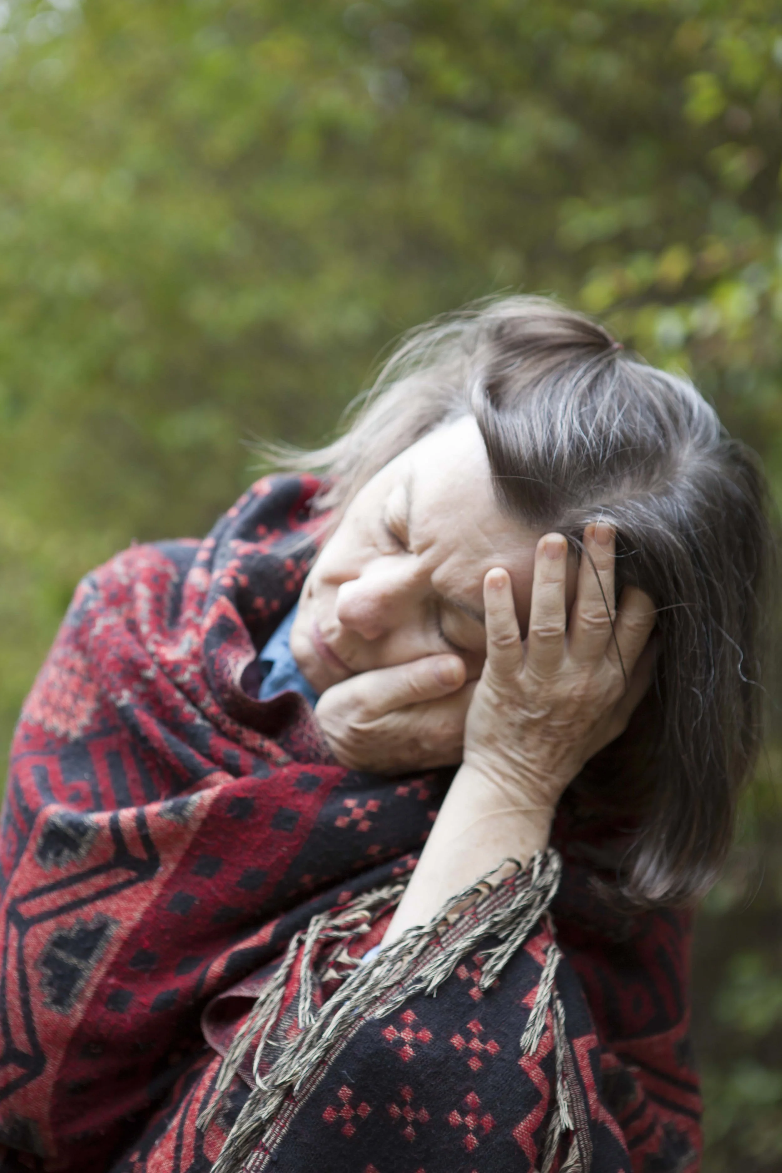An elderly woman with shoulder-length gray hair and closed eyes, resting her face on her hand while wearing a red and black patterned shawl outdoors with a blurry green background.