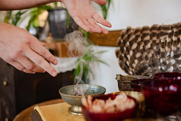 Hands reaching over a table with a steaming bowl, surrounded by bowls and a woven basket, in a cozy indoor setting.