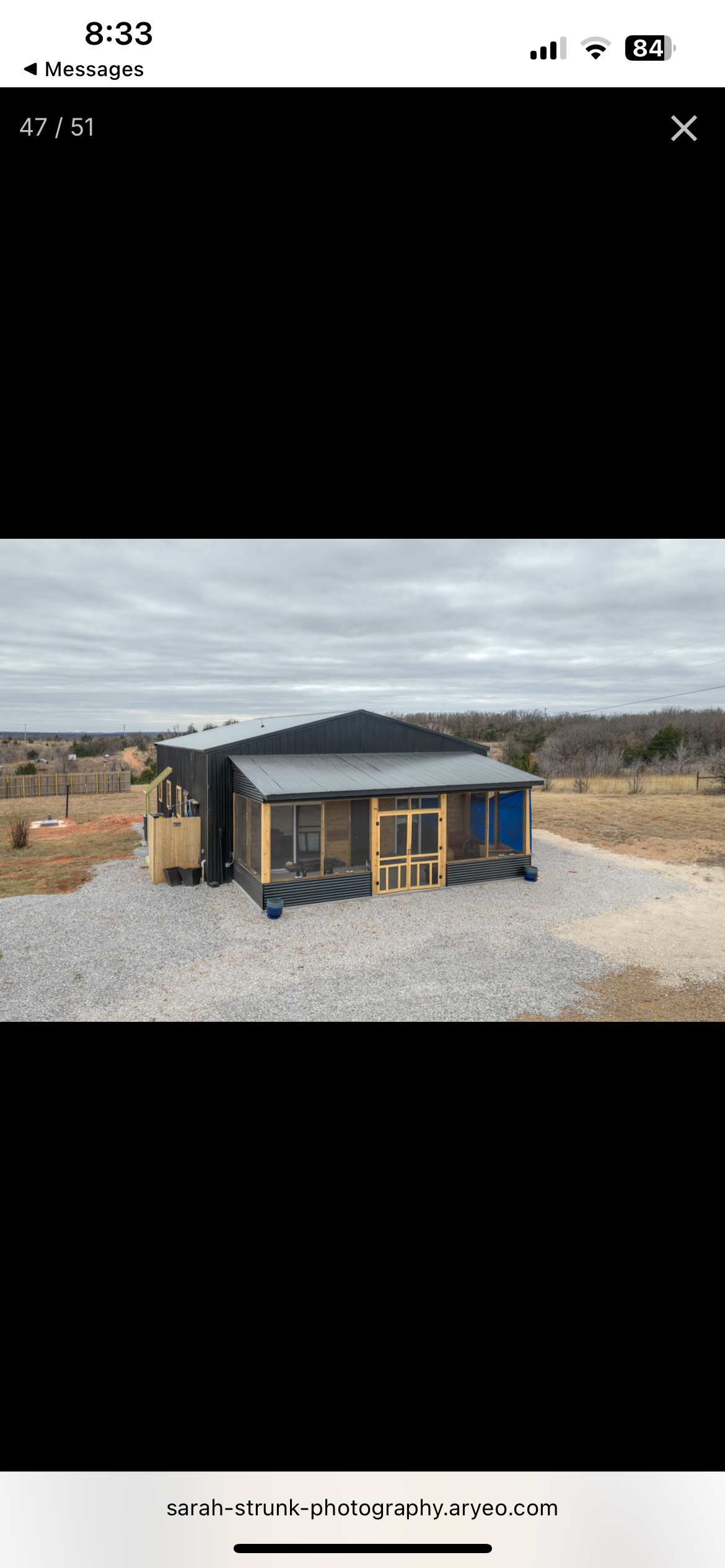 A small modern house with black exterior walls, a metal roof, and a screened porch, situated on a gravel lot with a fence and open land in the background under an overcast sky.