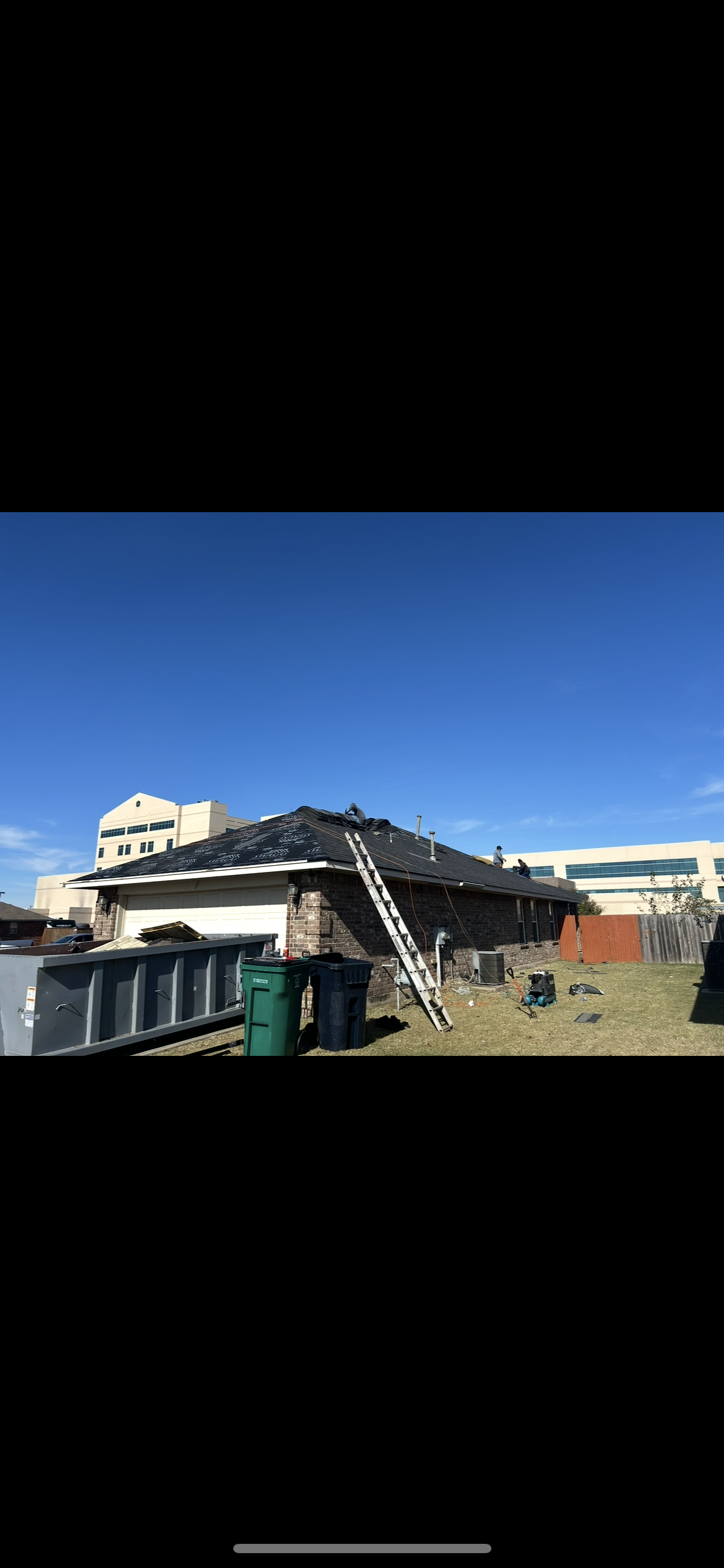 A house undergoing roof repairs with shingles removed, a ladder leaning against the roof, workers on the roof, and construction equipment in the yard under a clear blue sky.