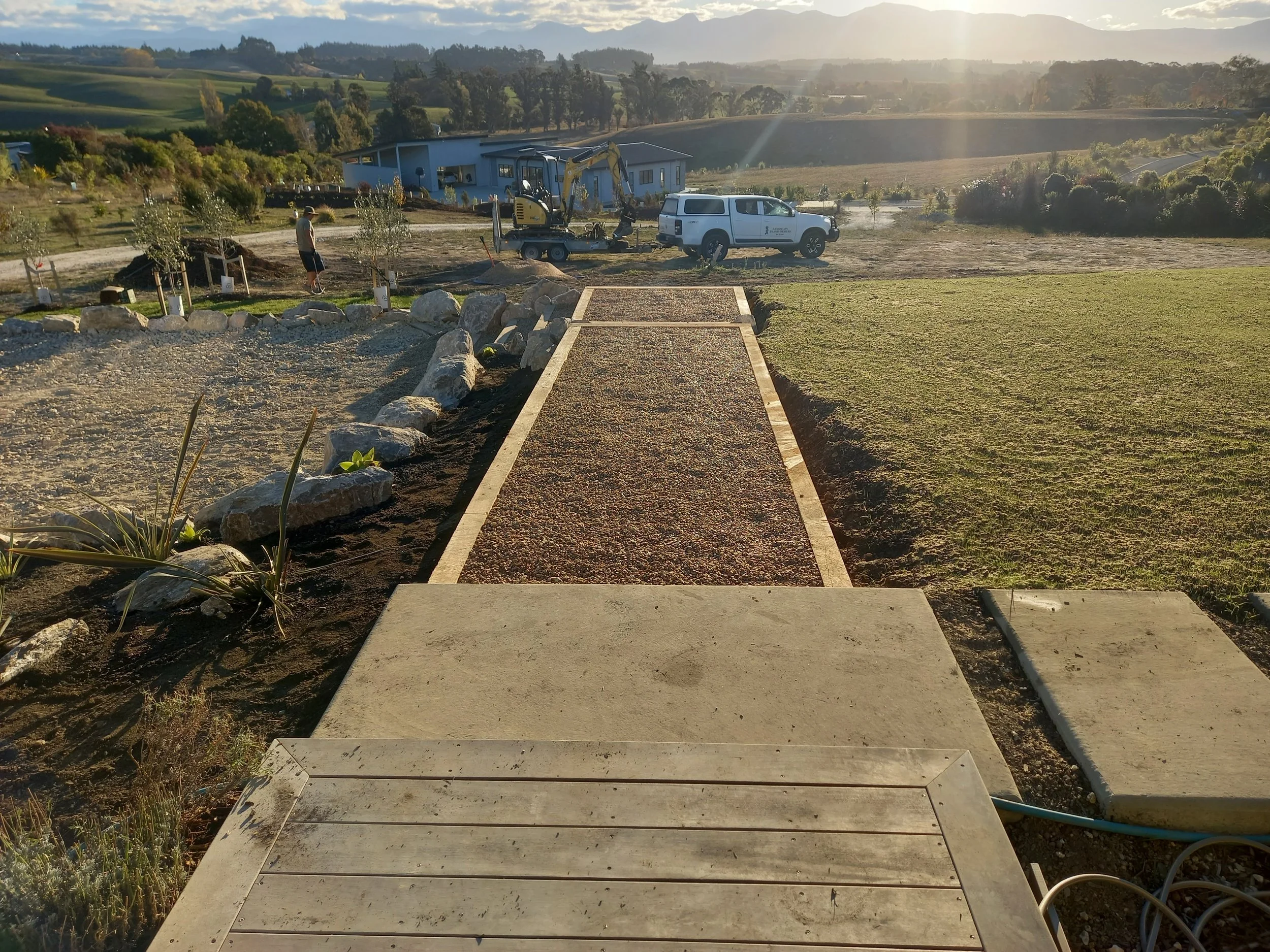 A construction site in a rural area with a pathway being built, surrounded by grass, rocks, and trees, with a white vehicle, construction equipment, and workers nearby, during sunset or sunrise.