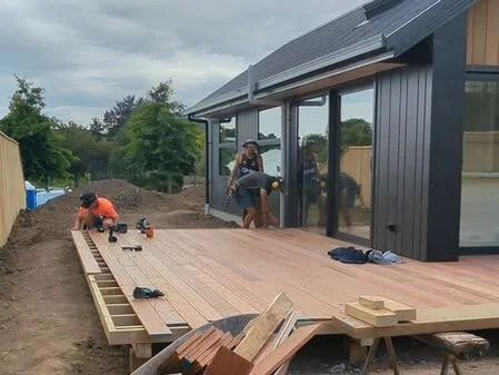 Two workers installing a wooden deck outside a modern house with large glass windows.